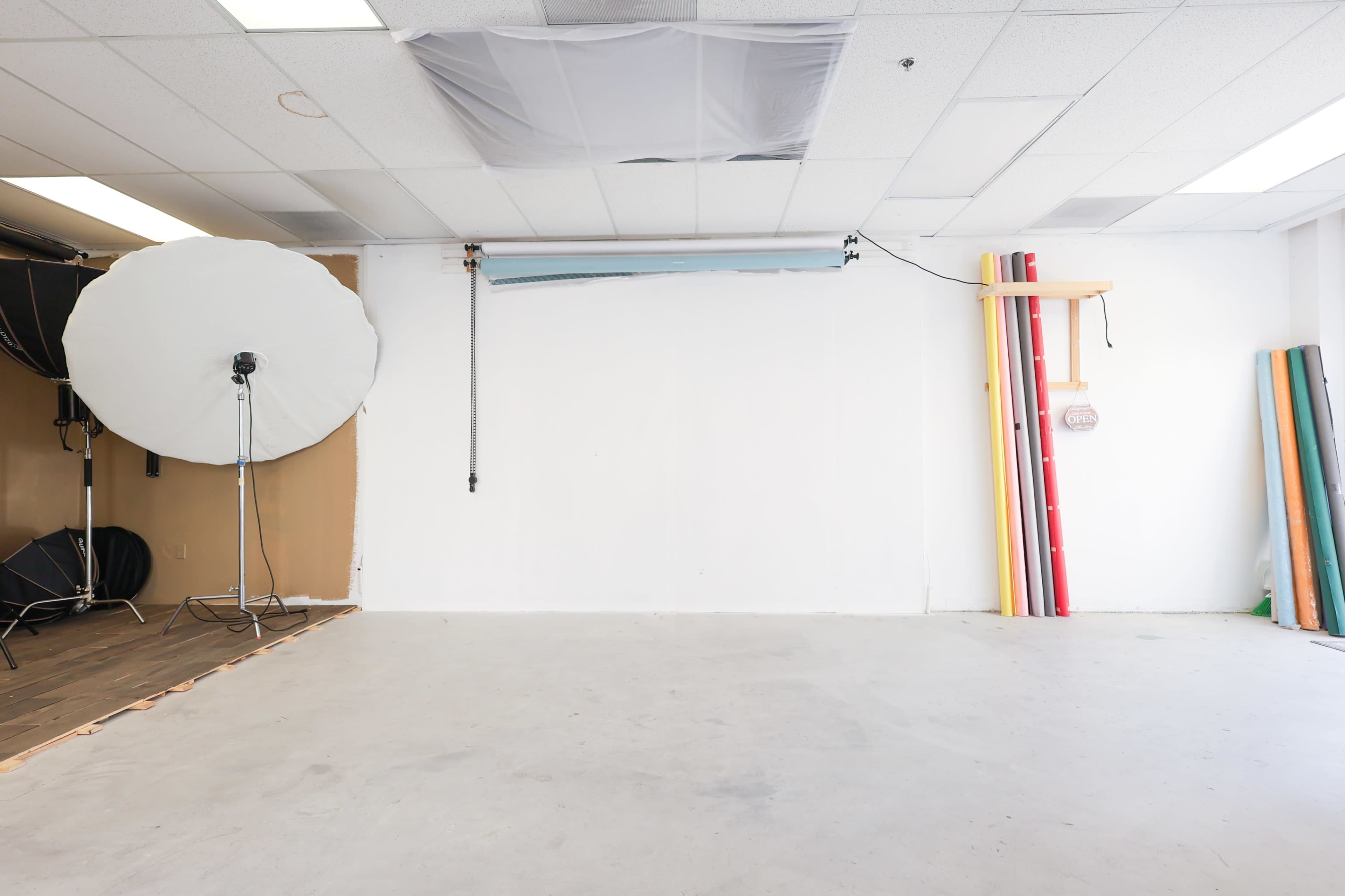 The image shows a spacious photography studio featuring a white backdrop, a large softbox light on a stand, and colorful backdrop rolls leaning against the wall.