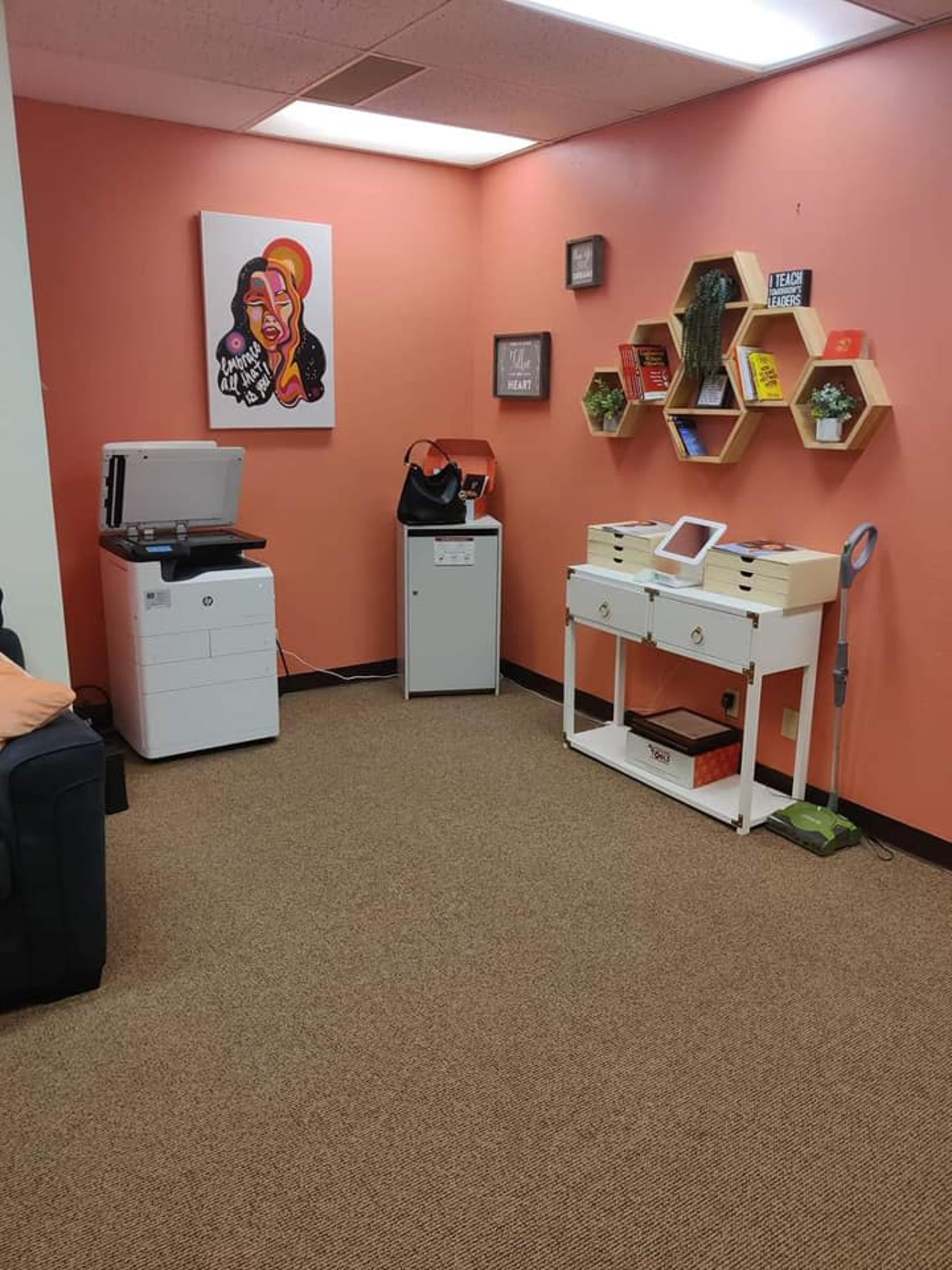 An office space with a printer, a small white table with a tablet, a filing cabinet, and a decorative wall display made of hexagonal shelves.