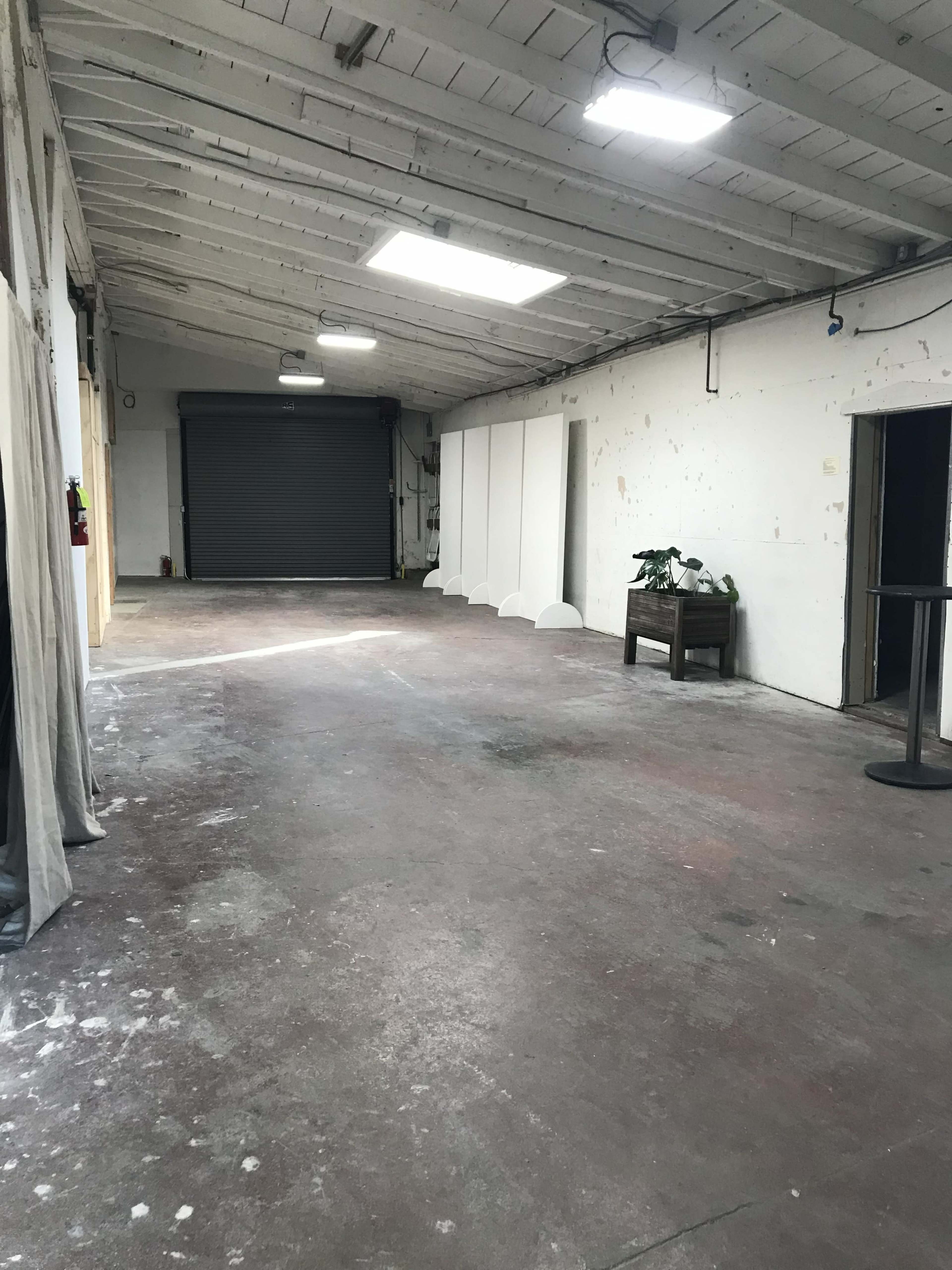 The image shows an empty industrial space with a concrete floor, partially lit by overhead skylights, featuring a few white partition panels and a single table with a plant.
