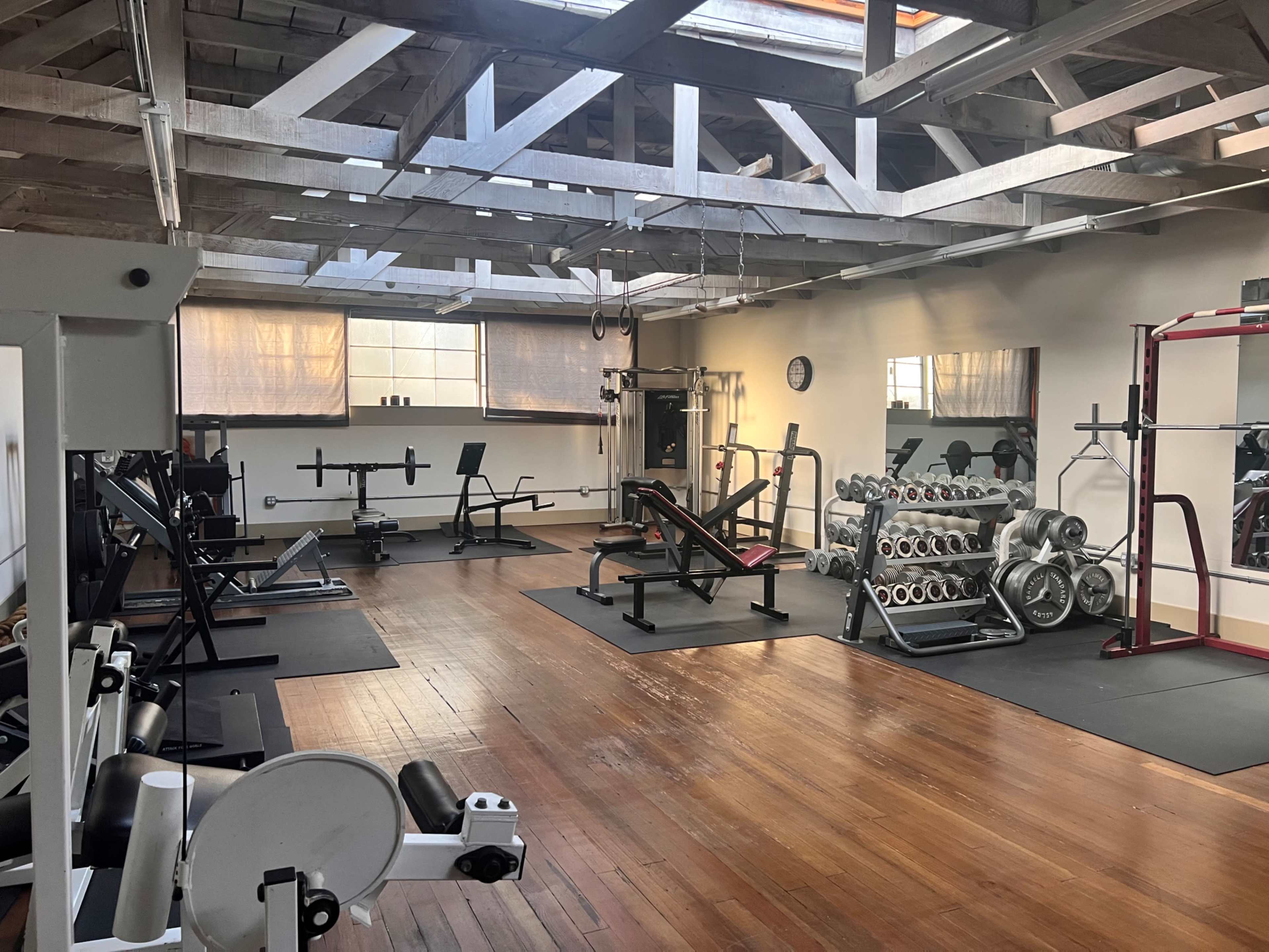 A spacious gym with various weightlifting equipment, benches, and racks, all arranged neatly on a wooden floor under a skylighted ceiling.