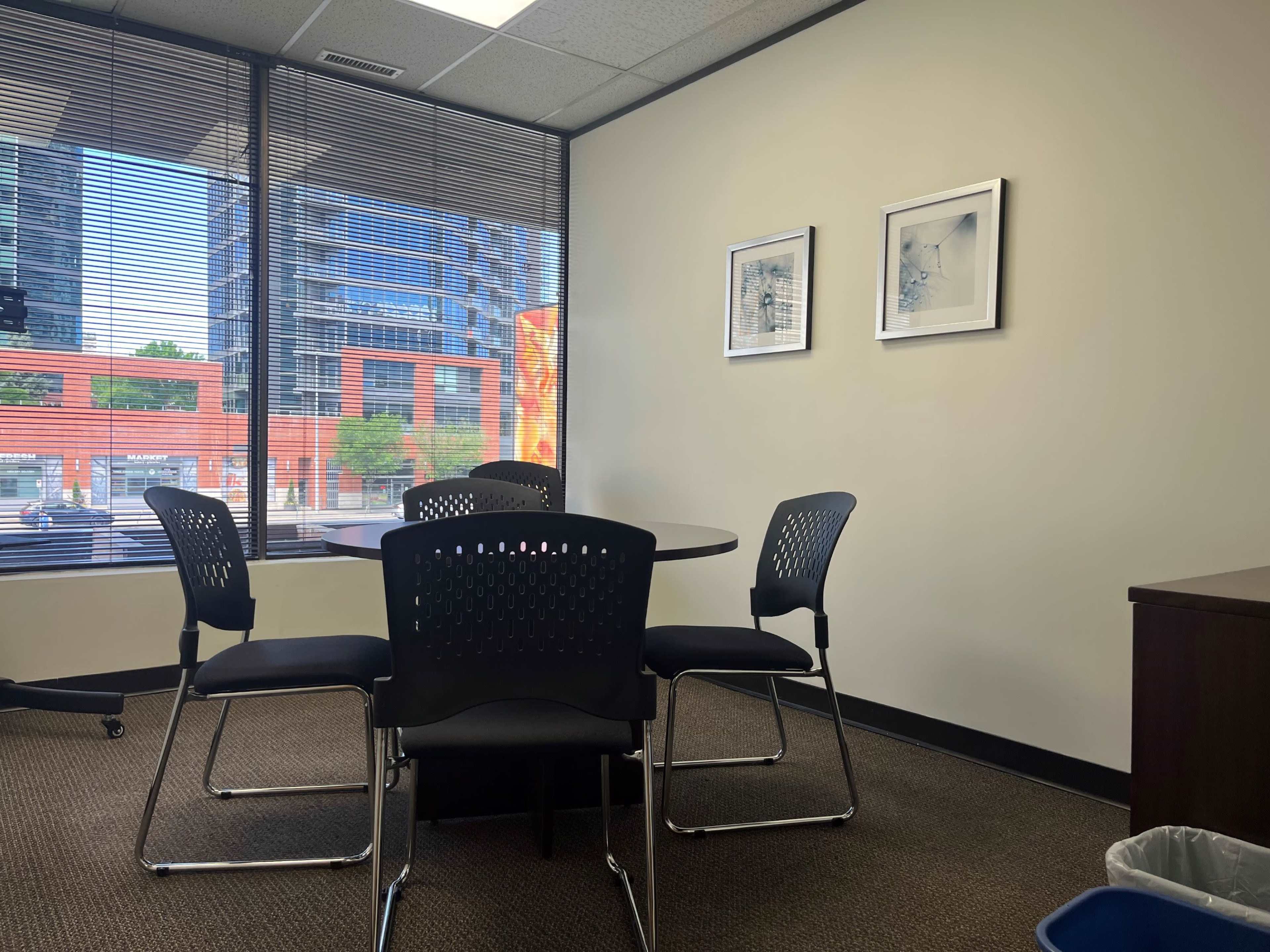 A small meeting room features a round table surrounded by four black chairs, with windows displaying a city view and abstract art on the walls.