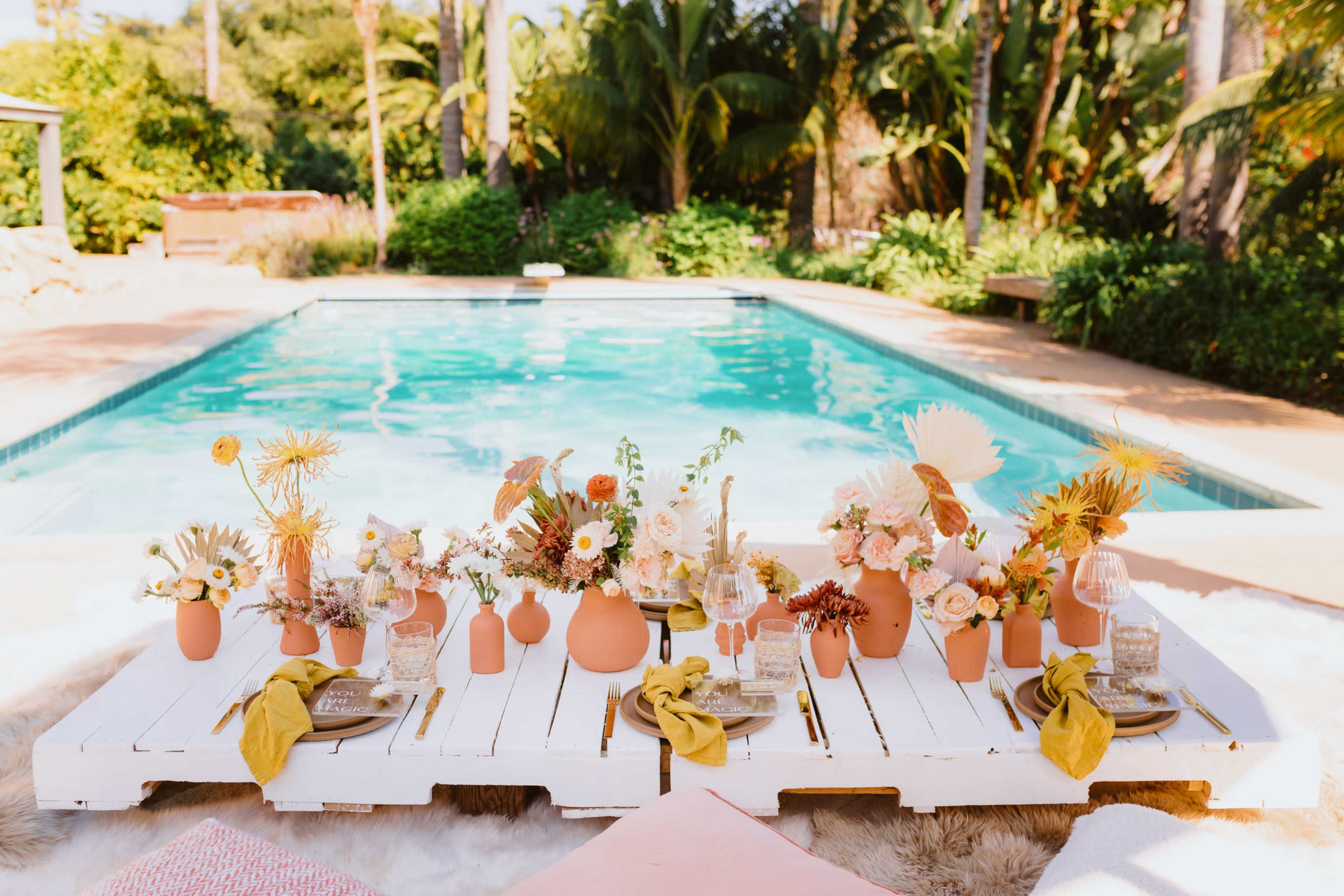 A styled outdoor dining setup featuring a wooden table adorned with colorful floral arrangements and tableware beside a swimming pool.