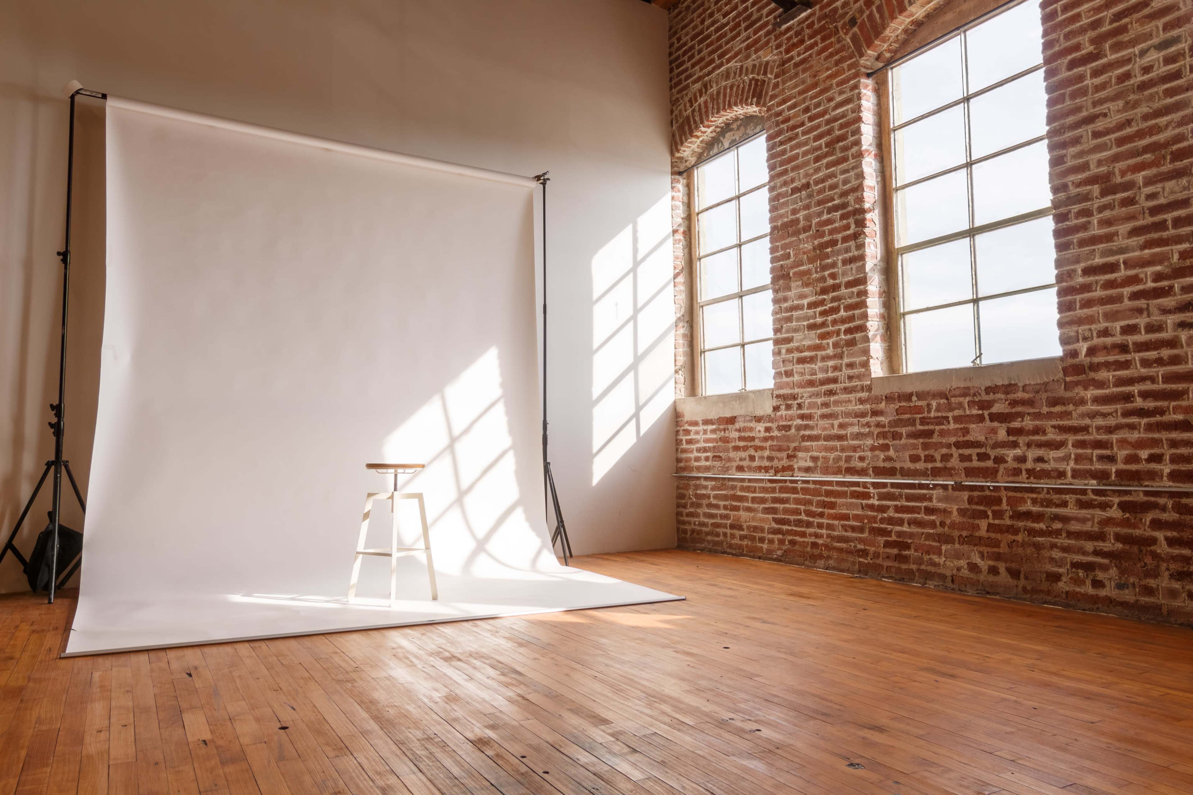 A photography studio features a blank backdrop with a wooden stool positioned in front of large windows that let in natural light.