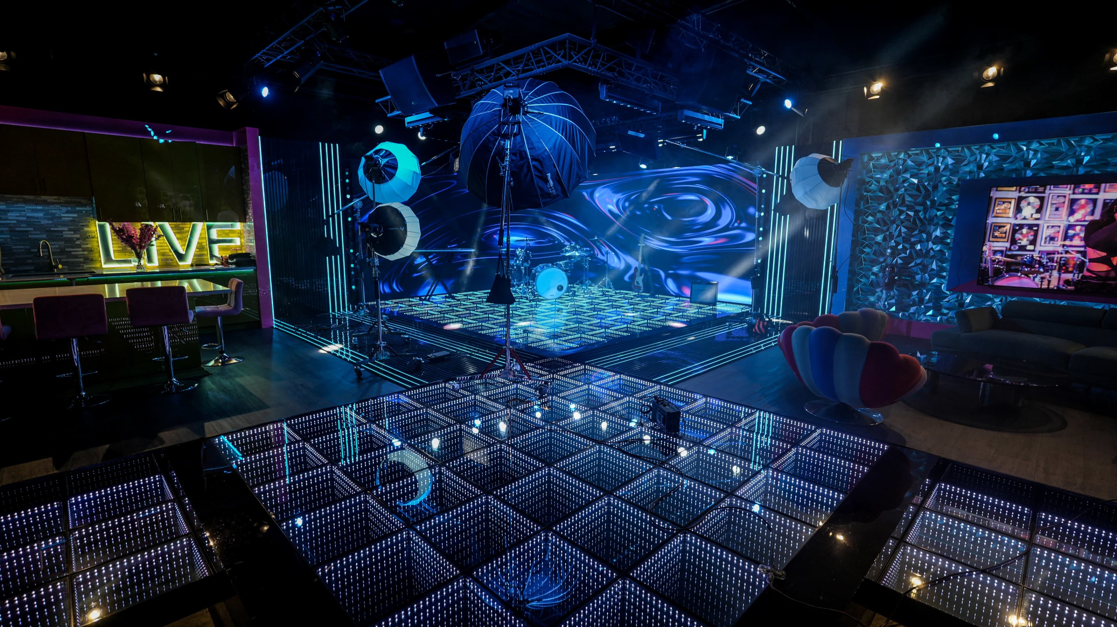 The image shows a vibrant television studio set with dynamic lighting, a patterned floor, and various colorful decorative elements.
