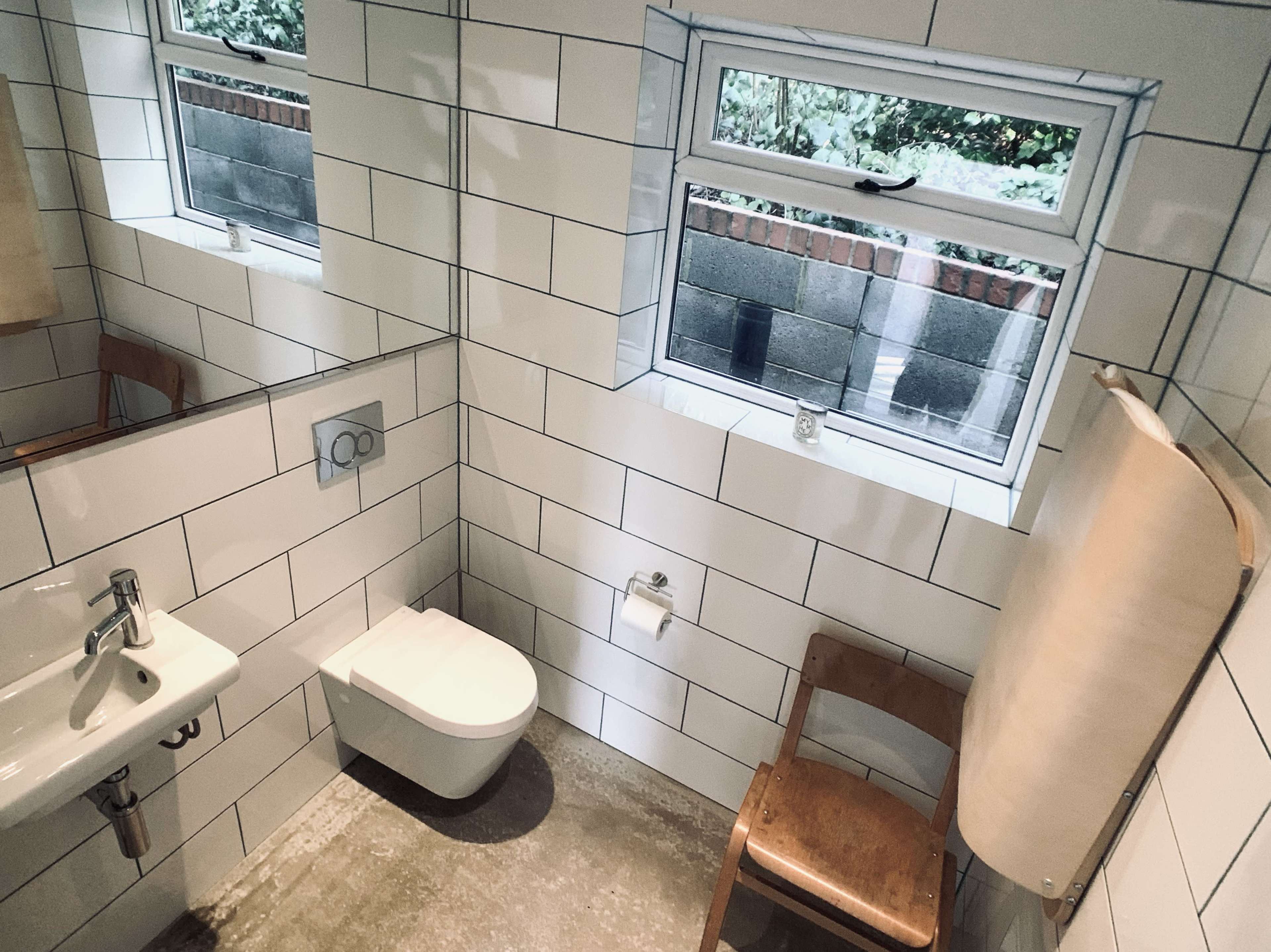 The image shows a modern bathroom featuring a wall-mounted toilet, a small sink, a wooden chair, and large tiled walls with two windows allowing natural light.