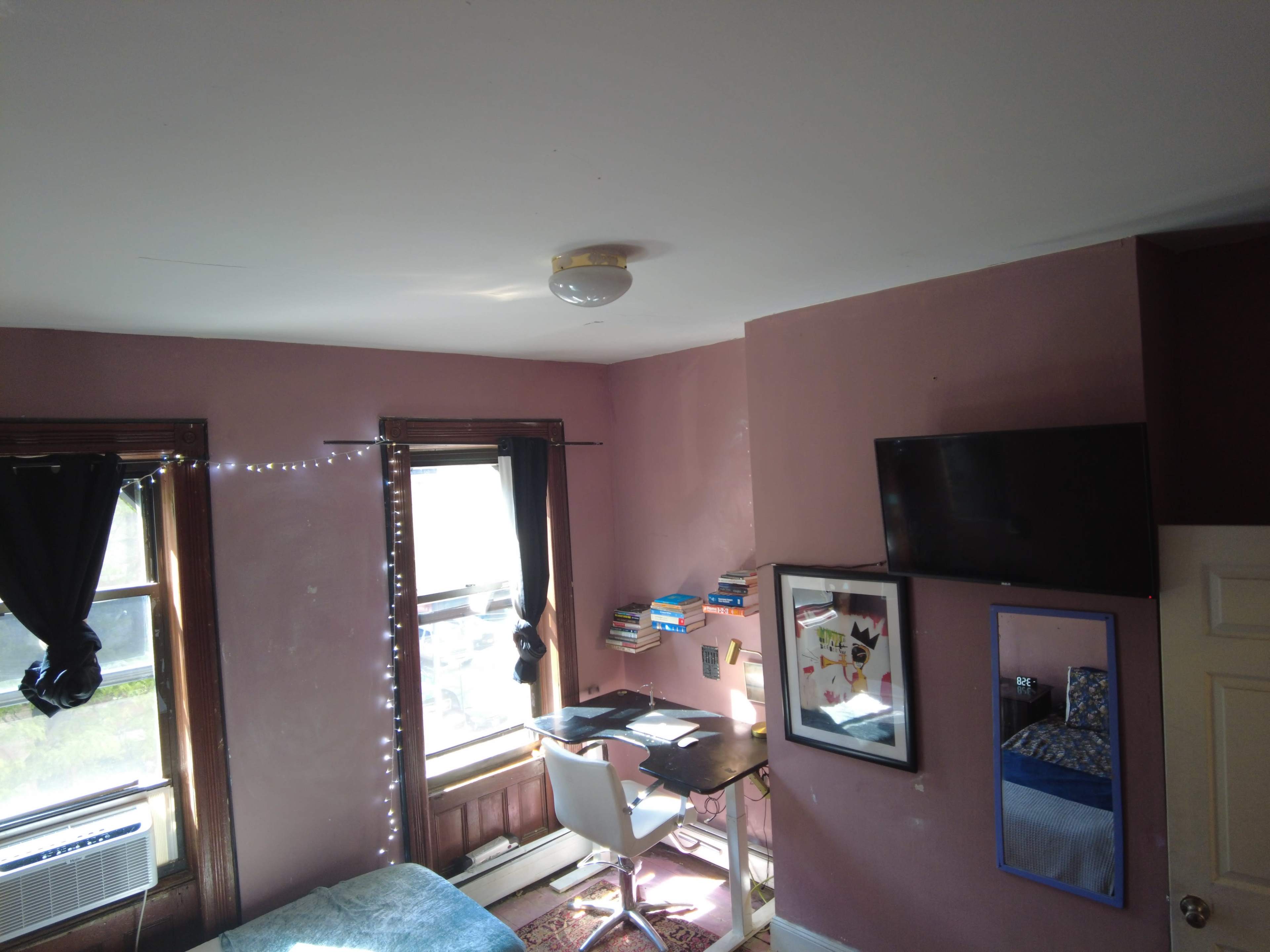 A small bedroom features a bed, a desk with a chair, a television on the wall, and a window with light curtains.