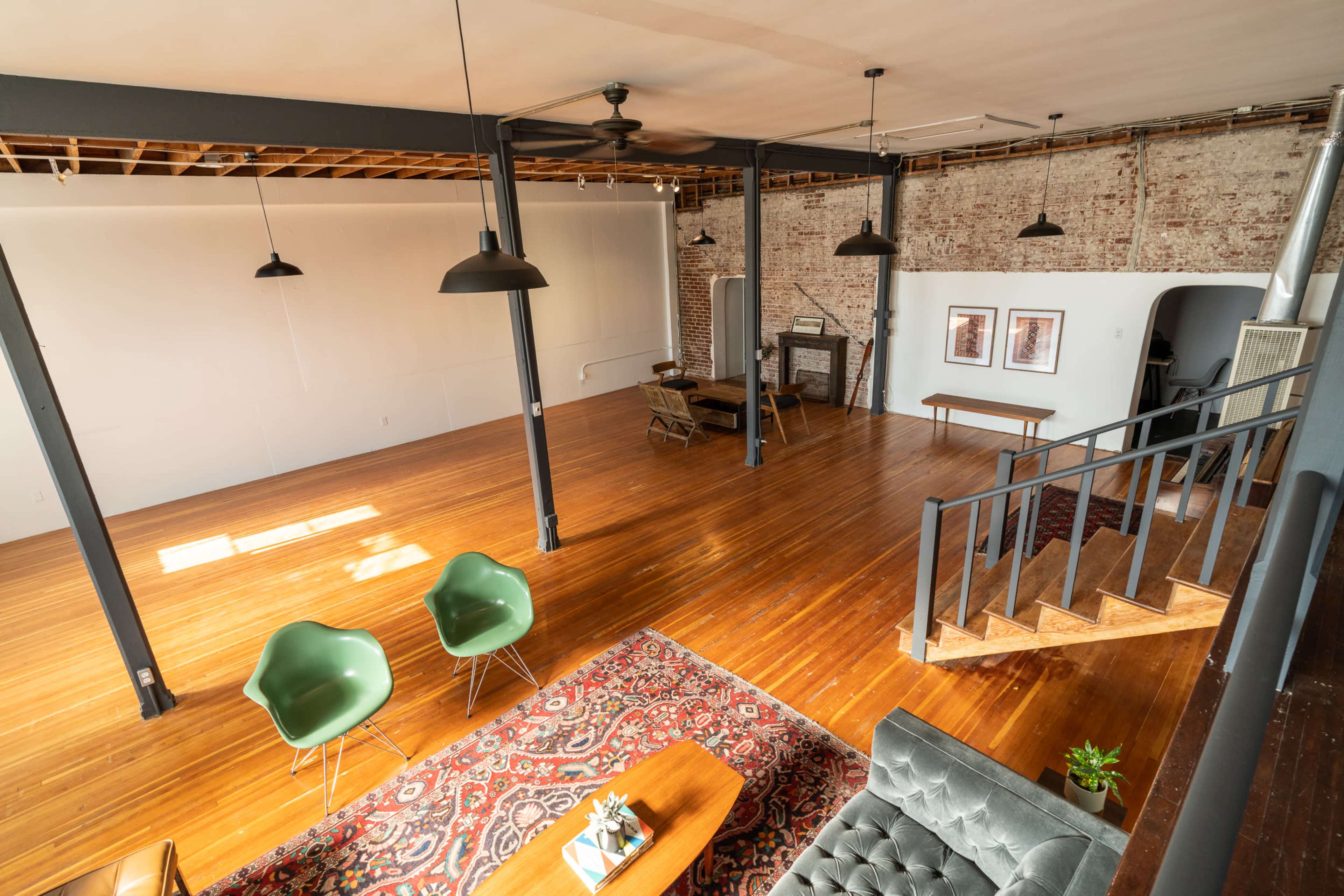 A spacious, open loft-style room with wooden floors, exposed brick walls, and modern furnishings, including green chairs and a gray sofa.