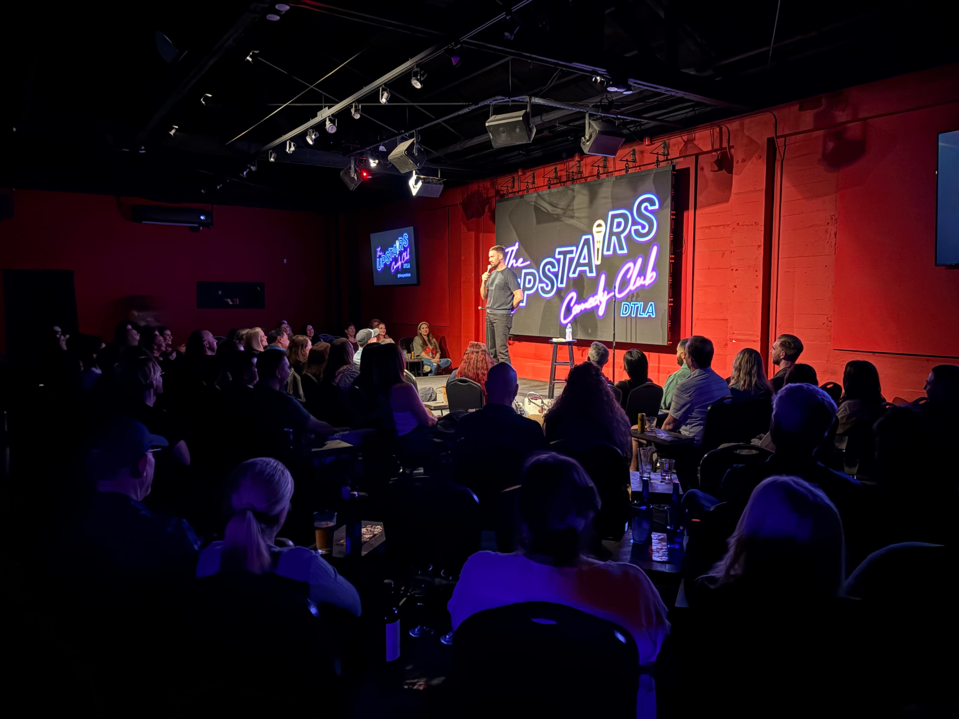 A comedian performs on stage at a comedy club filled with an audience seated at tables, illuminated by colorful lights.