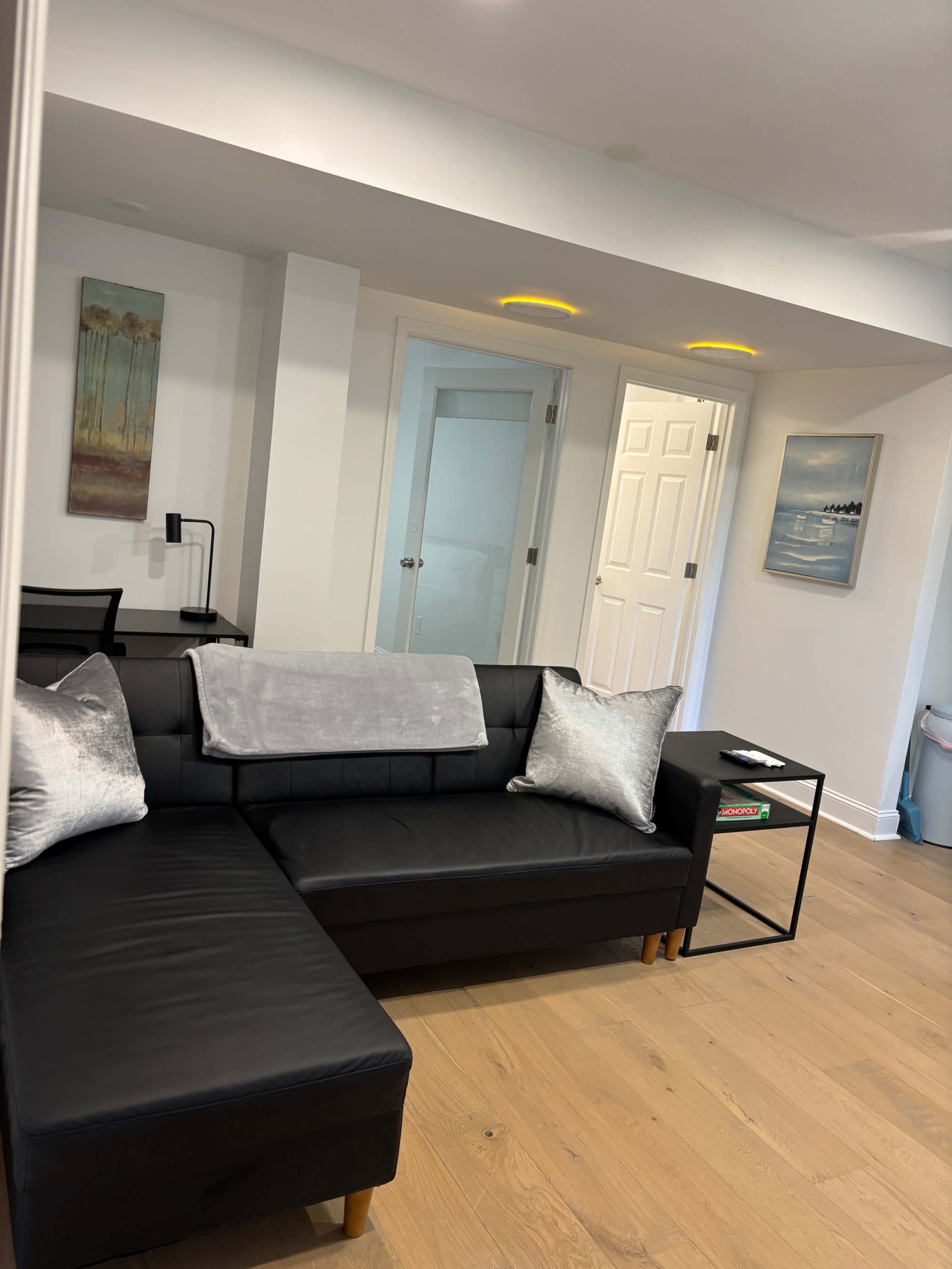The image shows a modern living room featuring a black sectional sofa, a small side table, and light-colored walls with artwork.