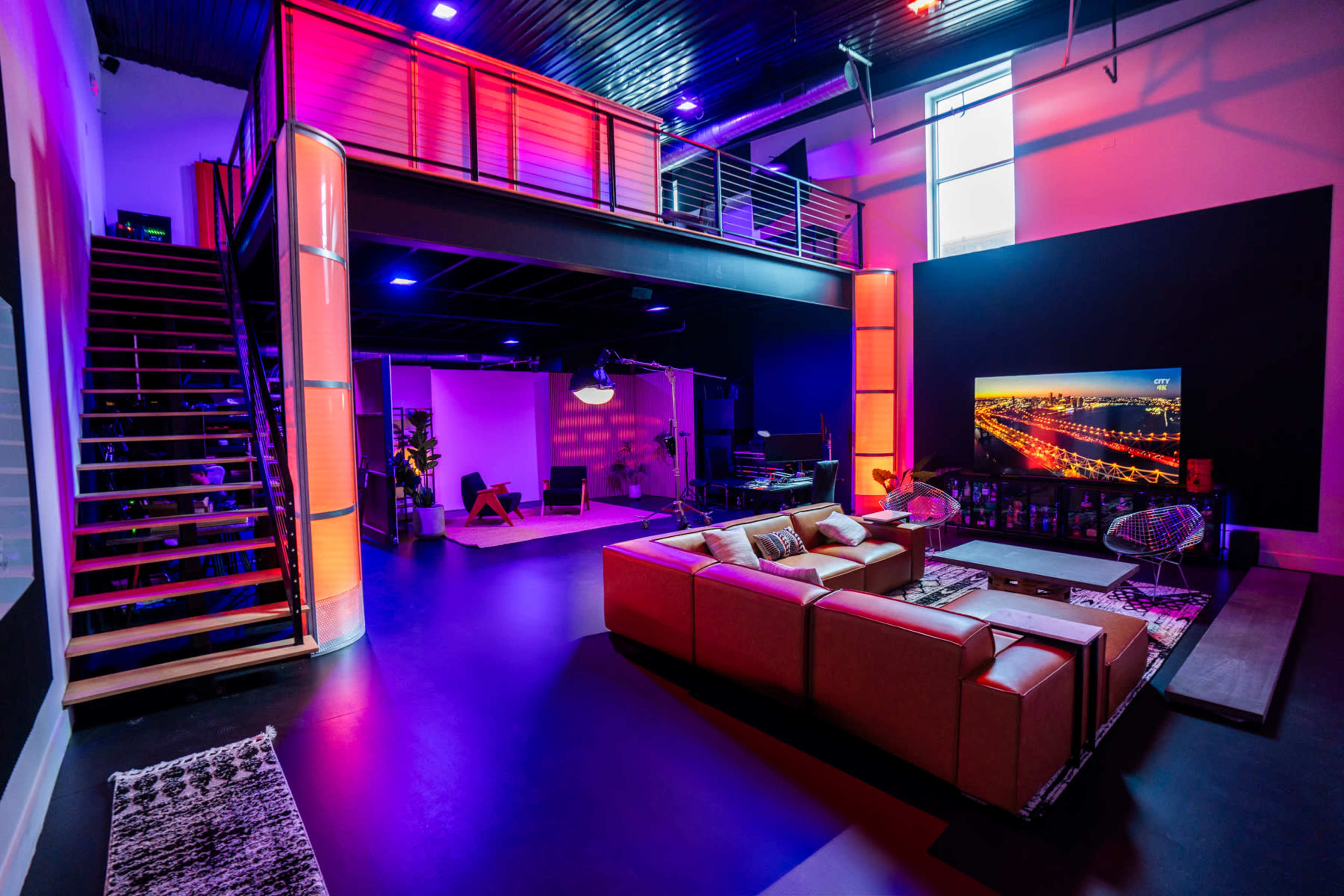 The image shows a modern interior space featuring a large open area with a staircase, seating arrangement, and a large TV screen, illuminated by colorful lighting.