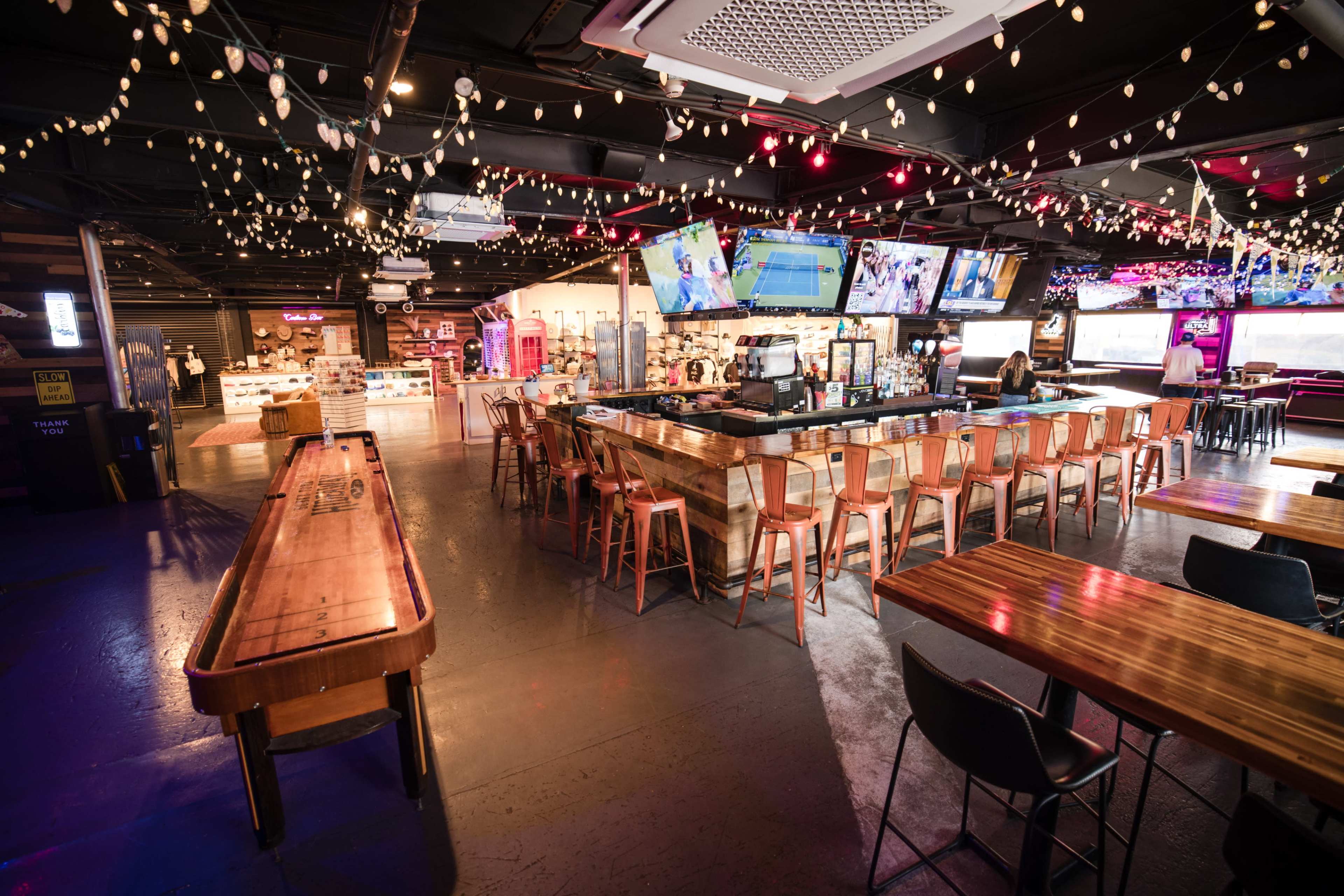 The image shows a spacious, modern bar and entertainment area with illuminated decorations, multiple screens displaying sports, and a shuffleboard table in the foreground.
