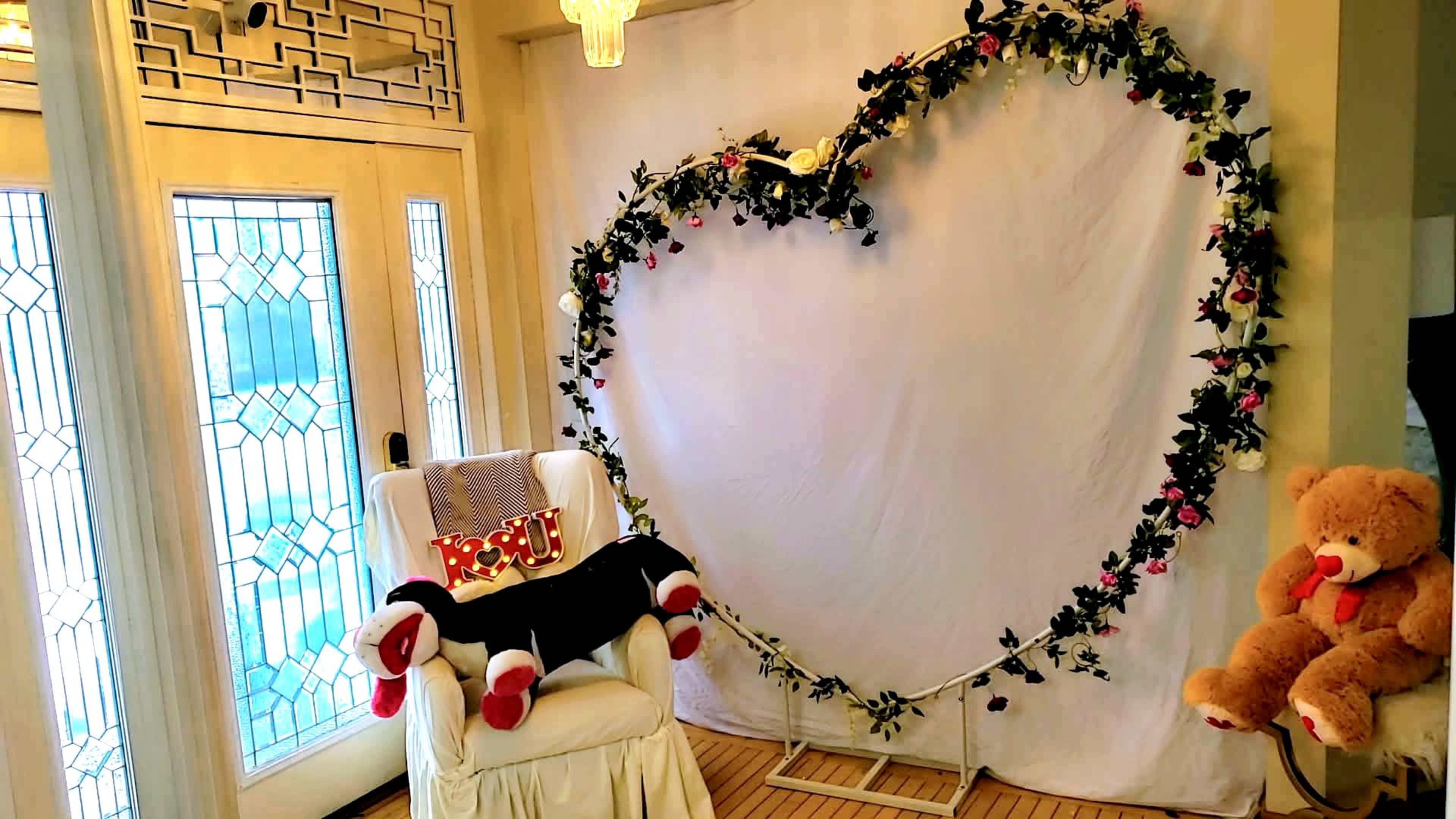 The scene features a white backdrop shaped like a heart, decorated with greenery and flowers, alongside a plush chair and two teddy bears.