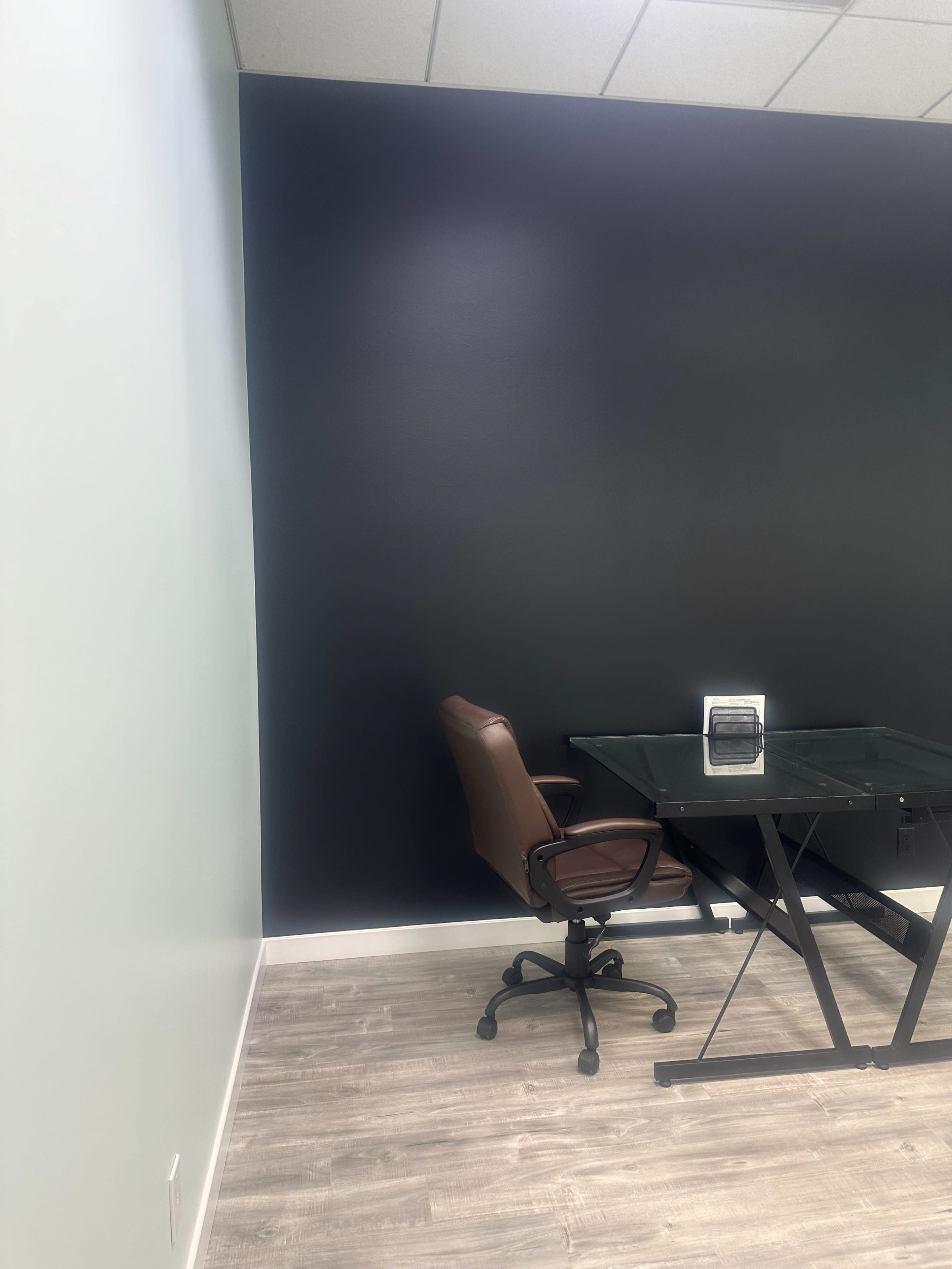 An office space with a brown chair and a glass desk against a black wall.