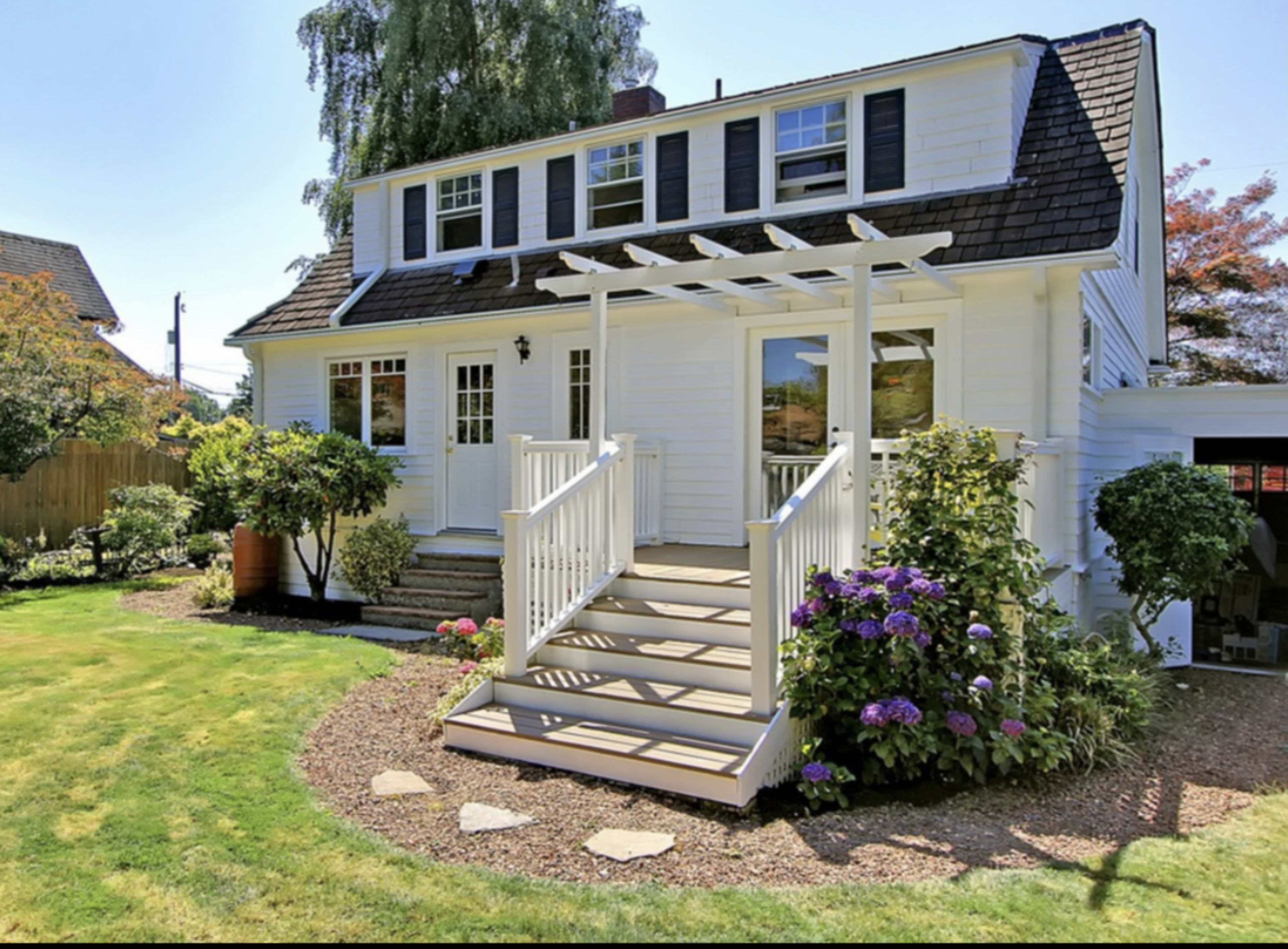 A two-story white house with a gabled roof features a front porch with a railing and landscaped gardens.