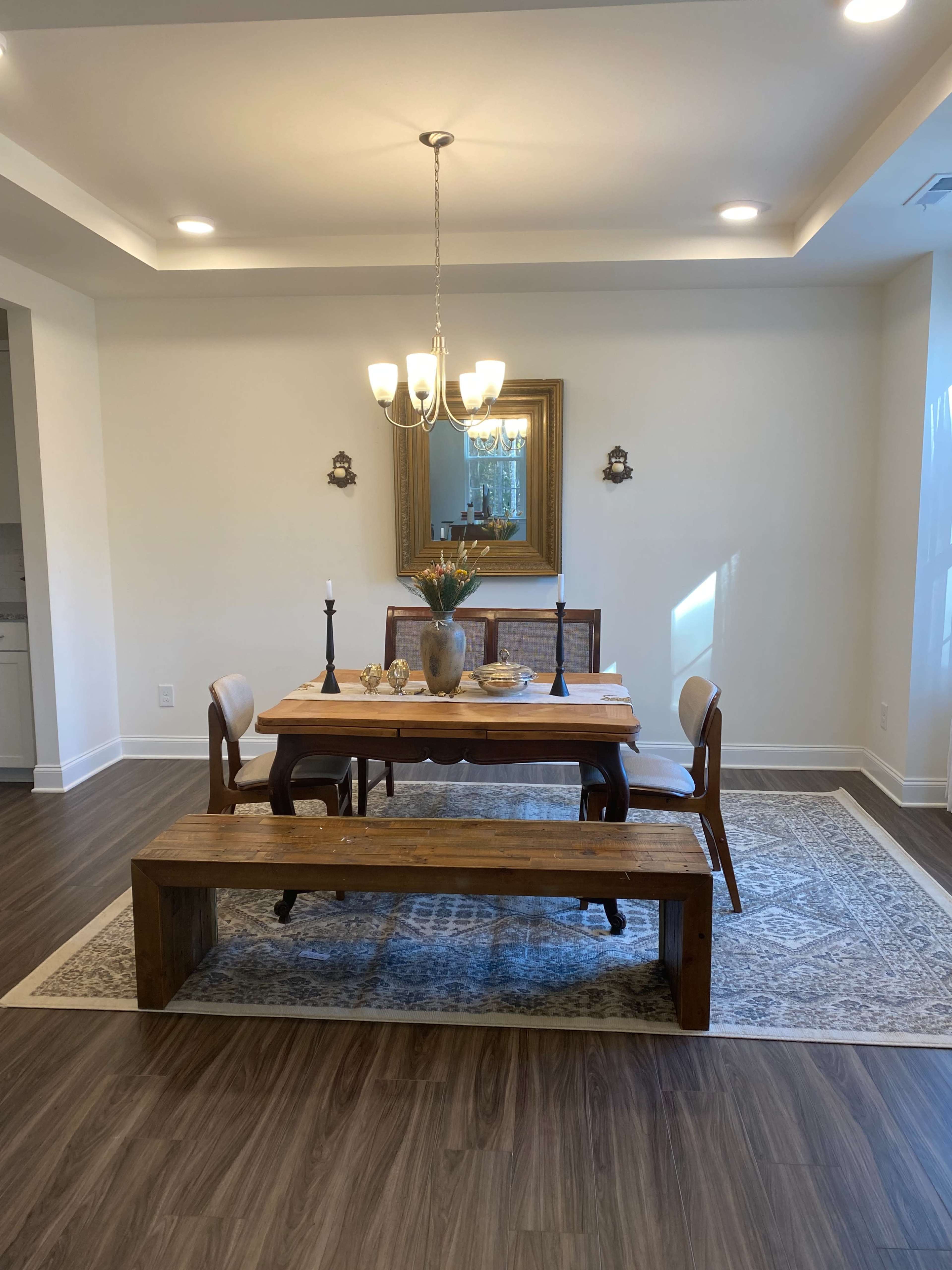 A dining area features a wooden table set for a meal, flanked by chairs and a long bench, under a chandelier and a large mirror.