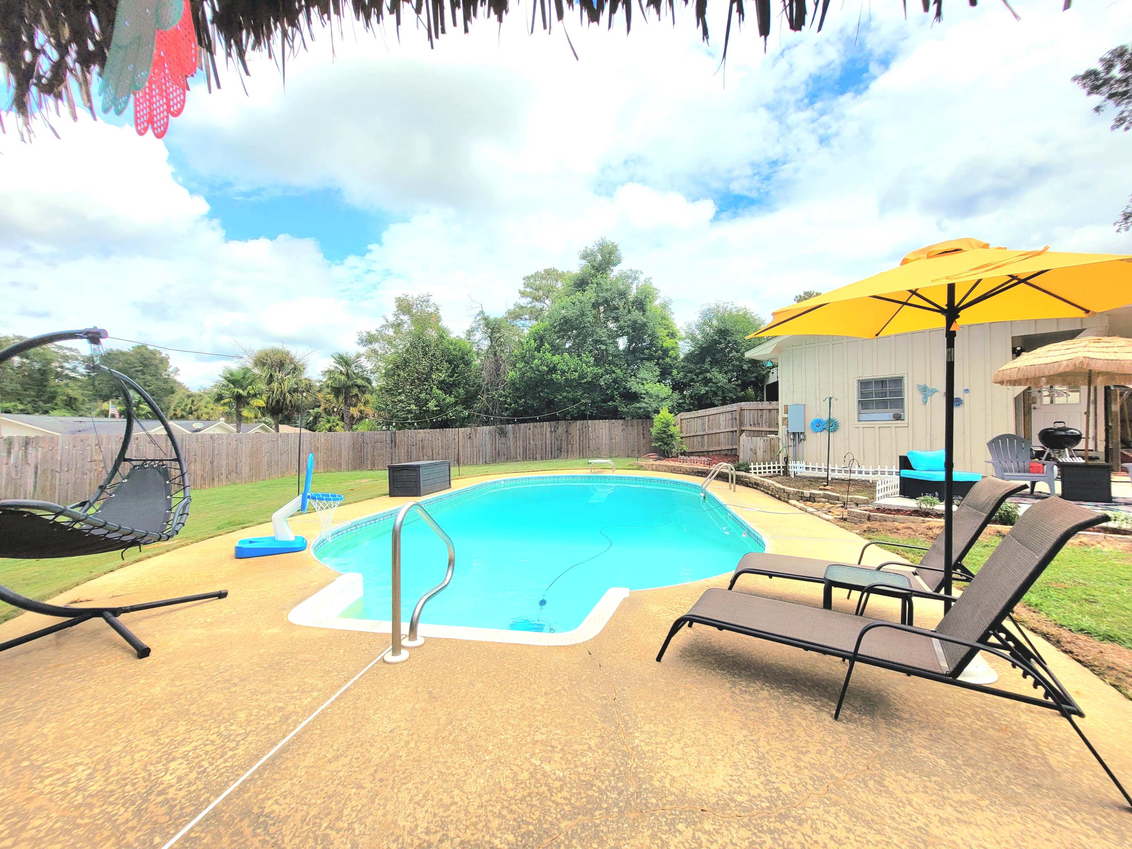 The image shows a backyard pool area with a clear blue pool, lounge chairs, and a yellow umbrella under a thatched structure.