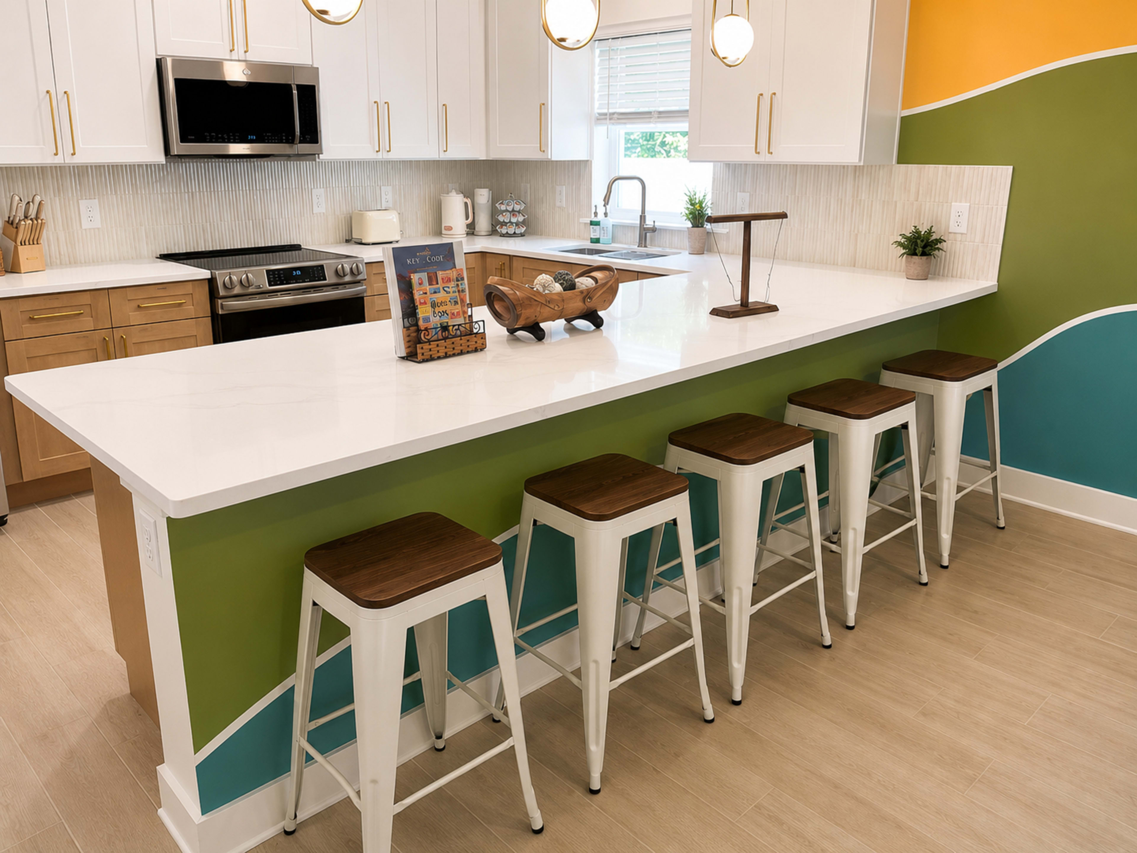 The image shows a modern kitchen with a long countertop, white cabinets, stainless steel appliances, and colorful accent walls.