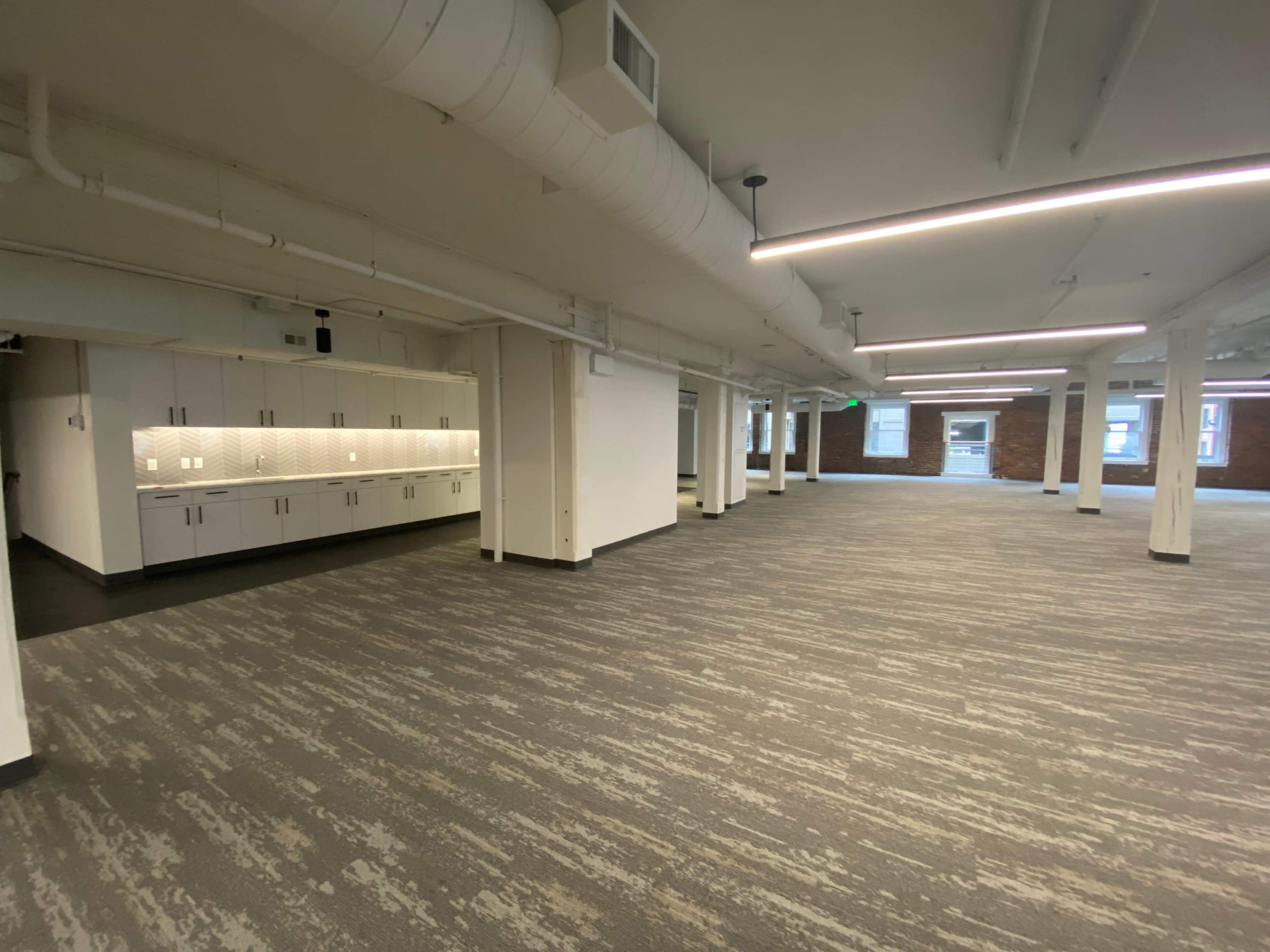 The image shows a large, empty office space with gray carpeting and white walls, featuring a kitchenette along one side.