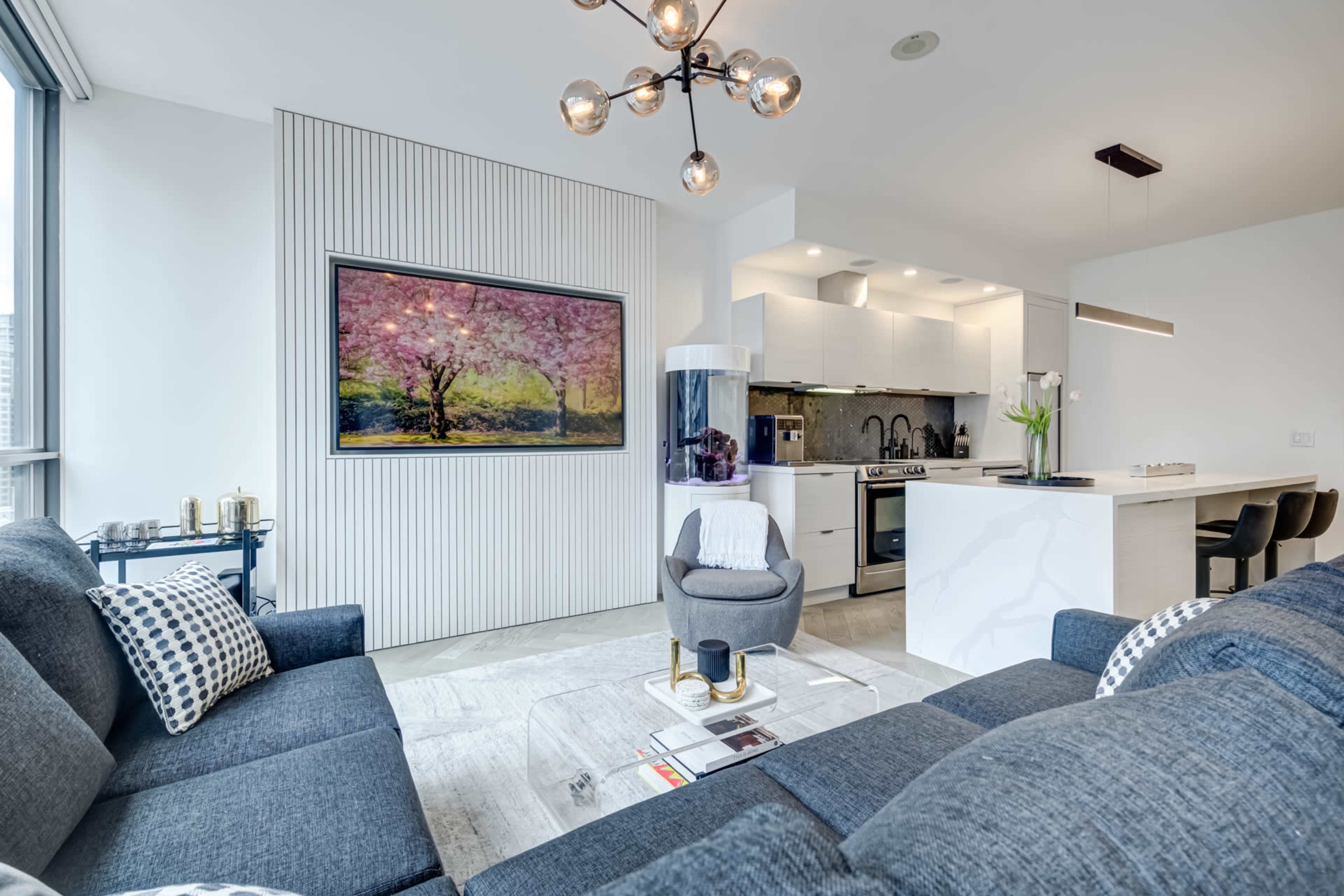 A modern living room features a dark gray sectional sofa, a glass coffee table, and a kitchen with white cabinets and a large wall-mounted painting.