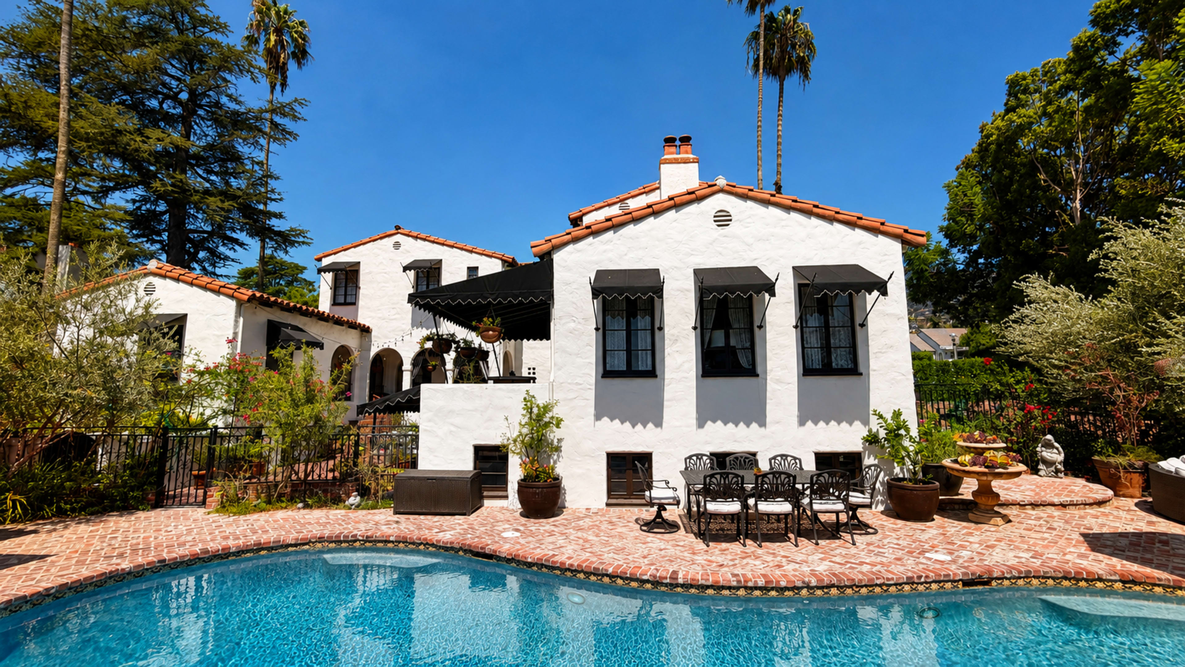 The image shows a white stucco house with a red tile roof beside a swimming pool, surrounded by lush greenery and patio furniture.