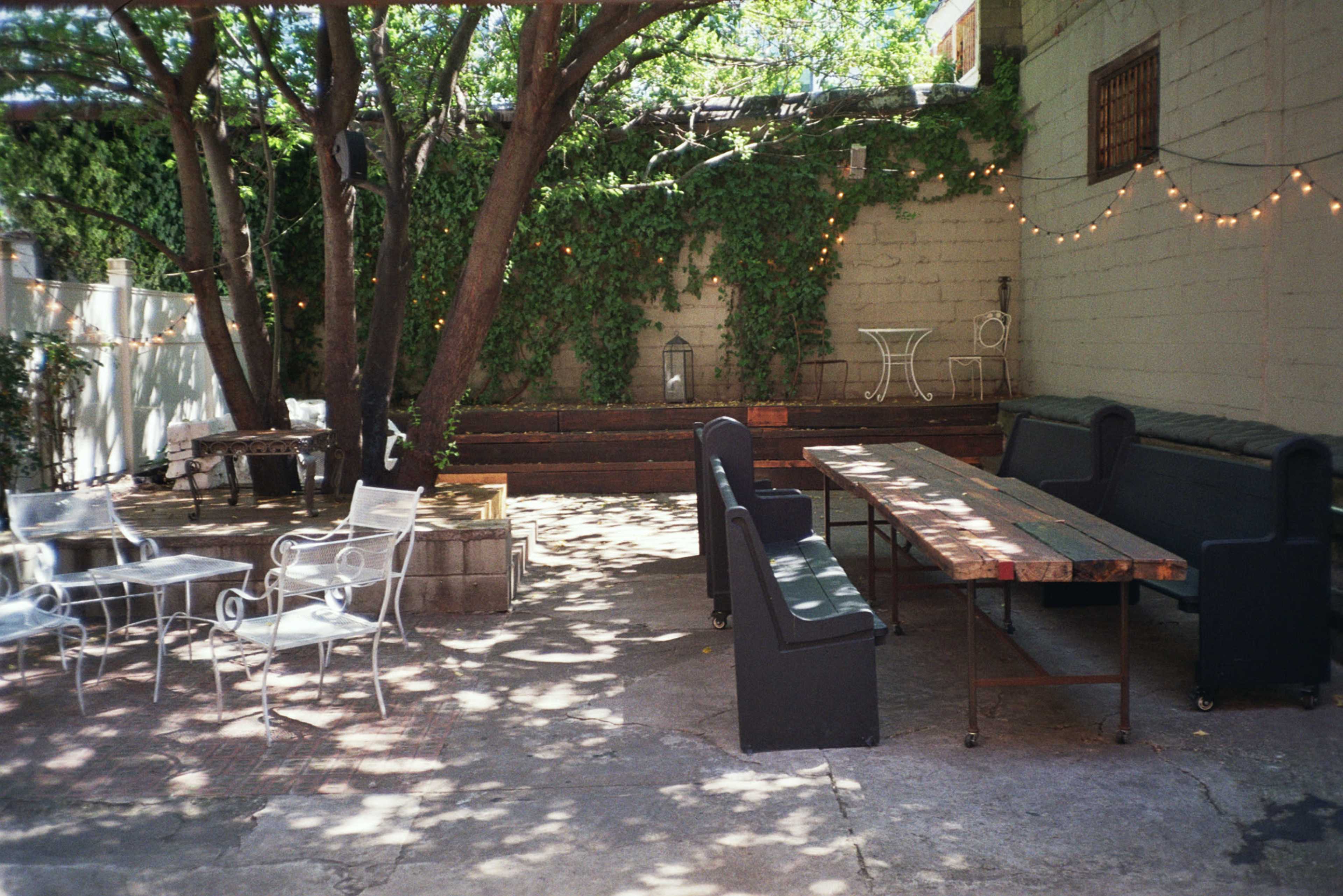 The image shows a shaded outdoor space with a long wooden table, several chairs, and decorative string lights hanging in a garden-like setting.