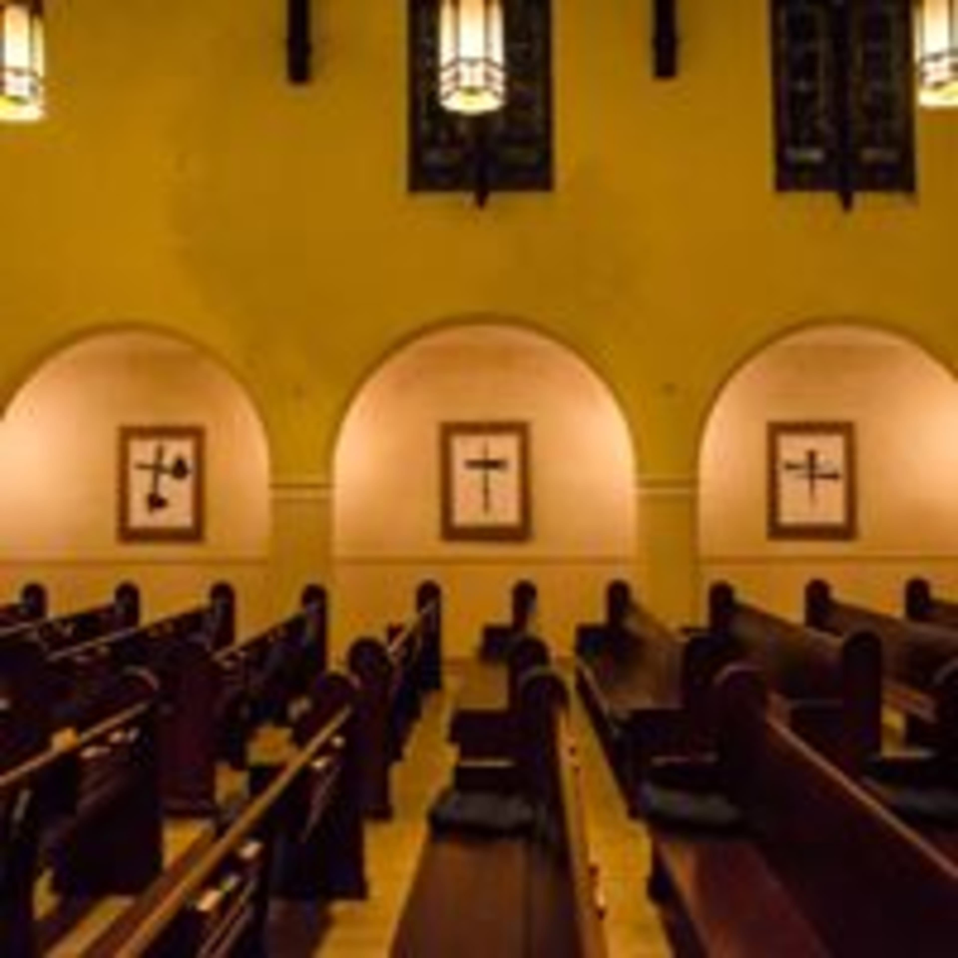 An interior of a church with wooden pews and framed crosses on the walls under arched openings.