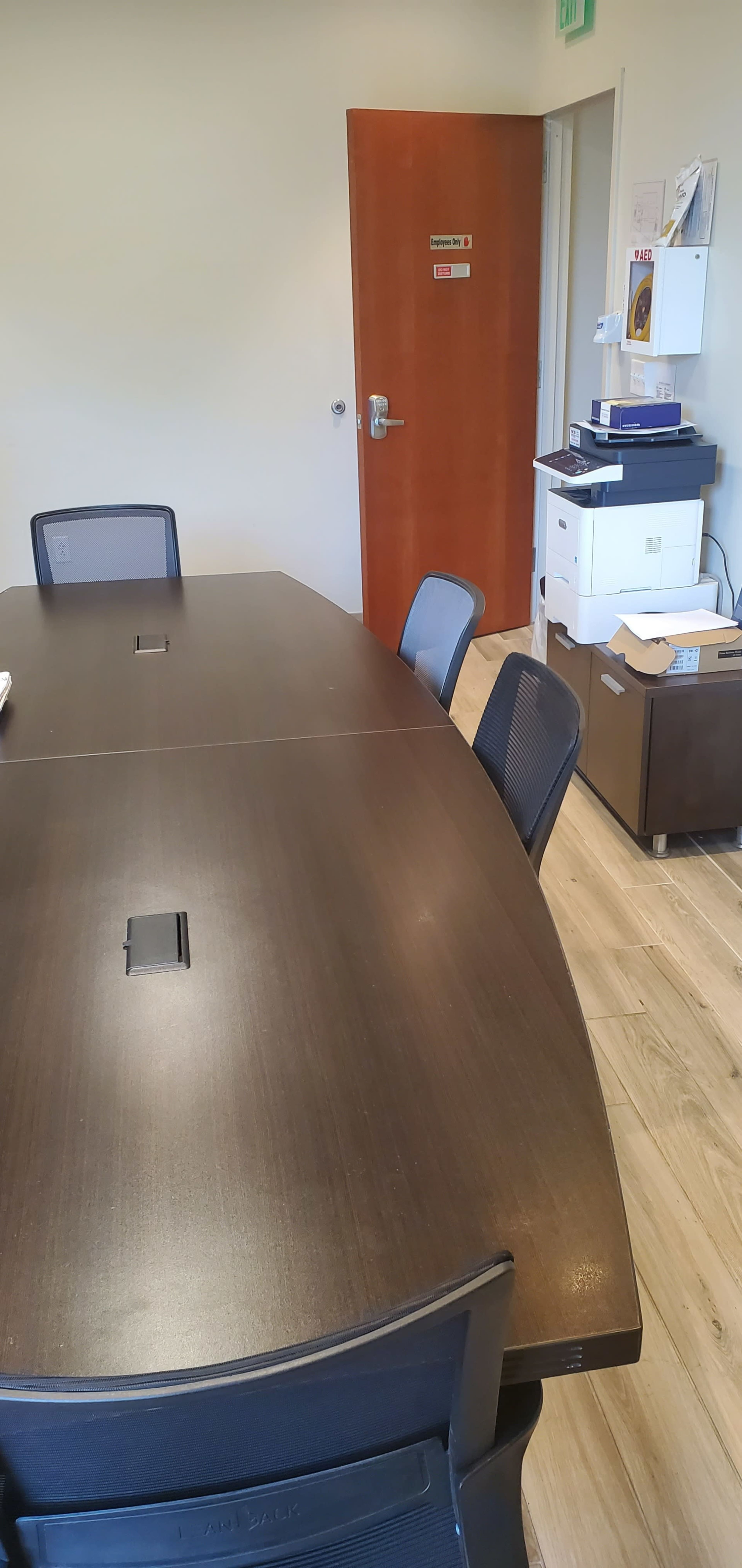 A conference room with a dark oval table and several black chairs is shown, along with a printer and shelving against the wall.