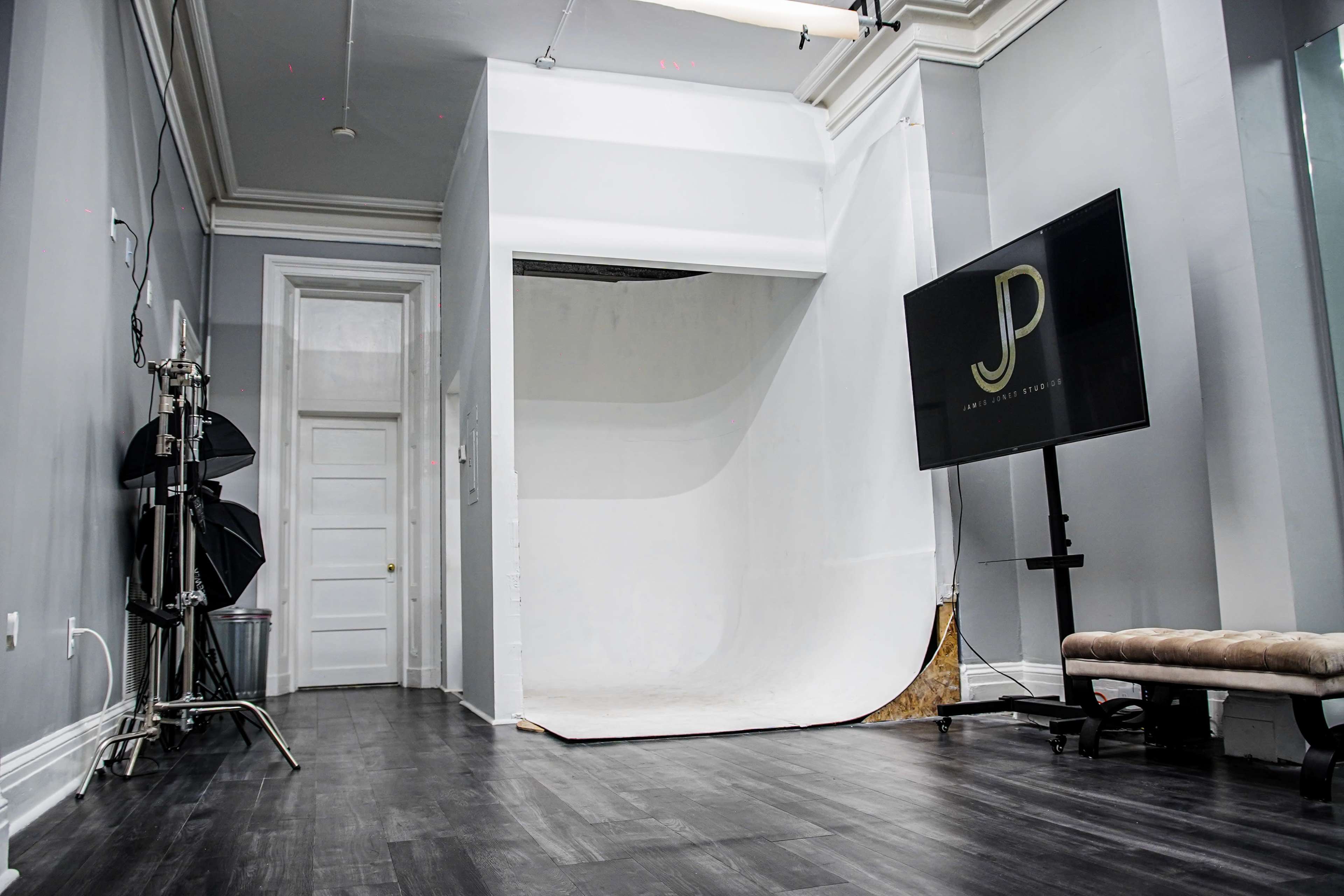 A spacious photography studio featuring a white backdrop, a television stand displaying a logo, and studio lighting equipment.