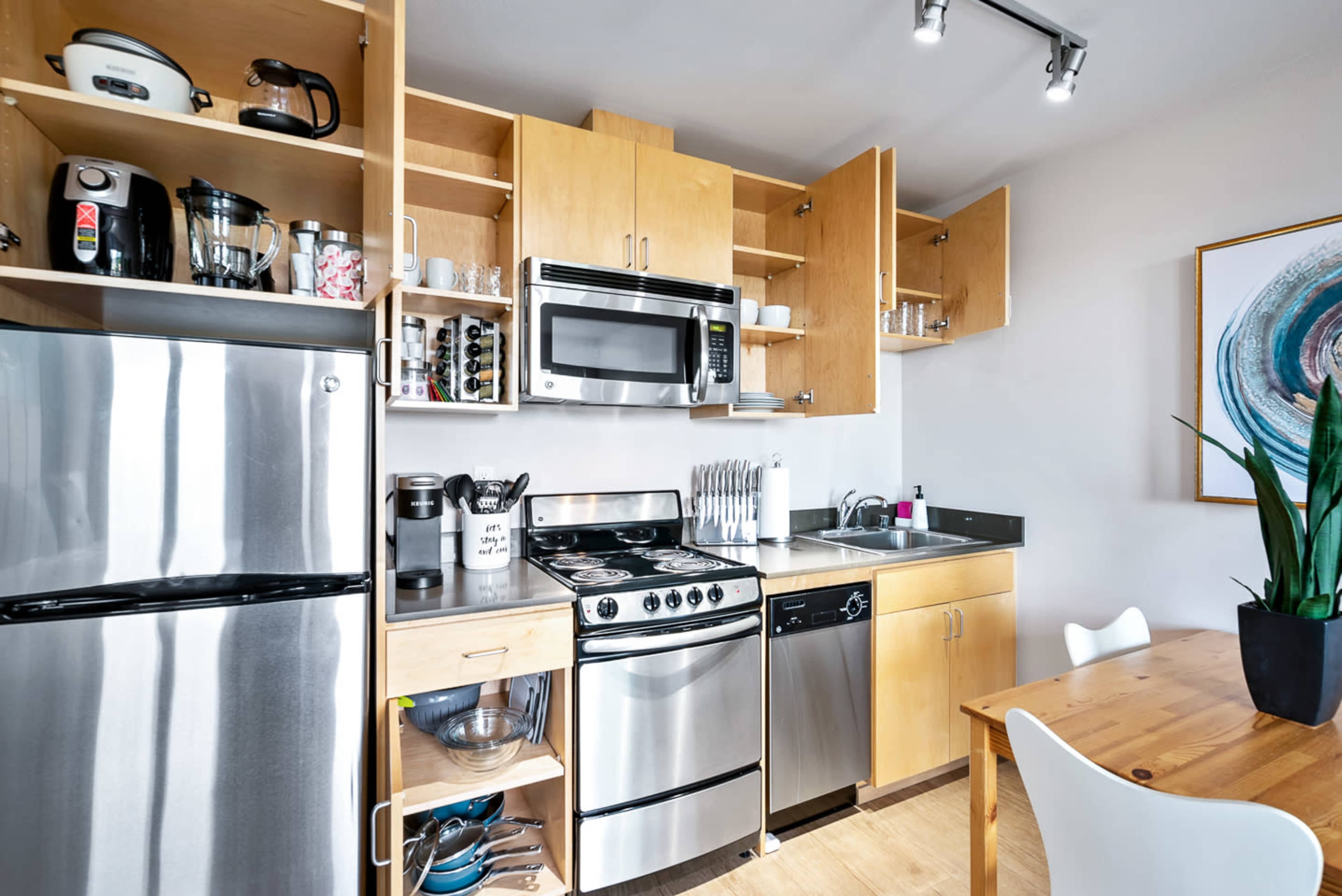 The image shows a compact kitchen featuring stainless steel appliances, wooden cabinetry, and a small dining table.
