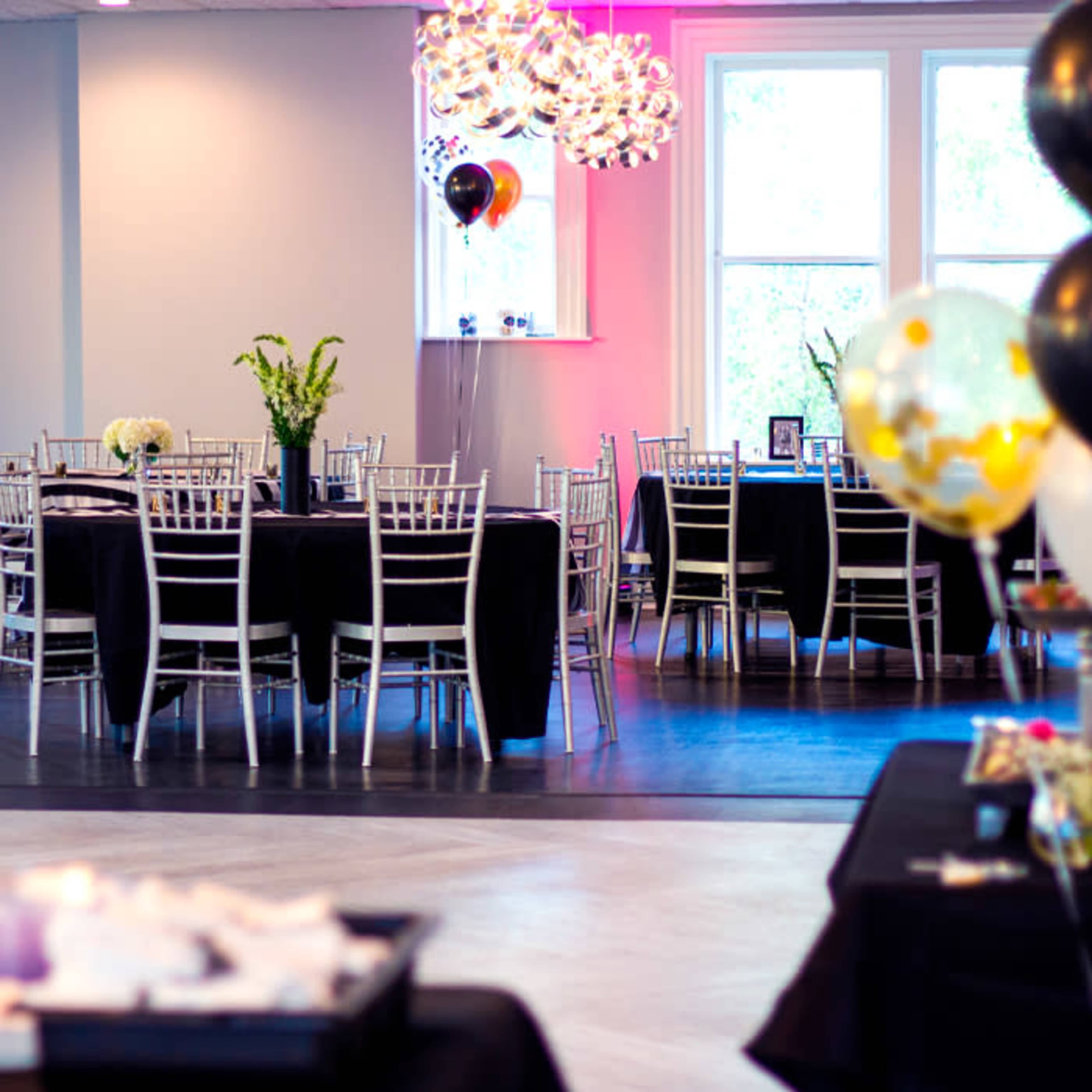 A room set up for a event features several floor tables with black cloths, silver chairs, decorative balloons, and a modern chandelier.