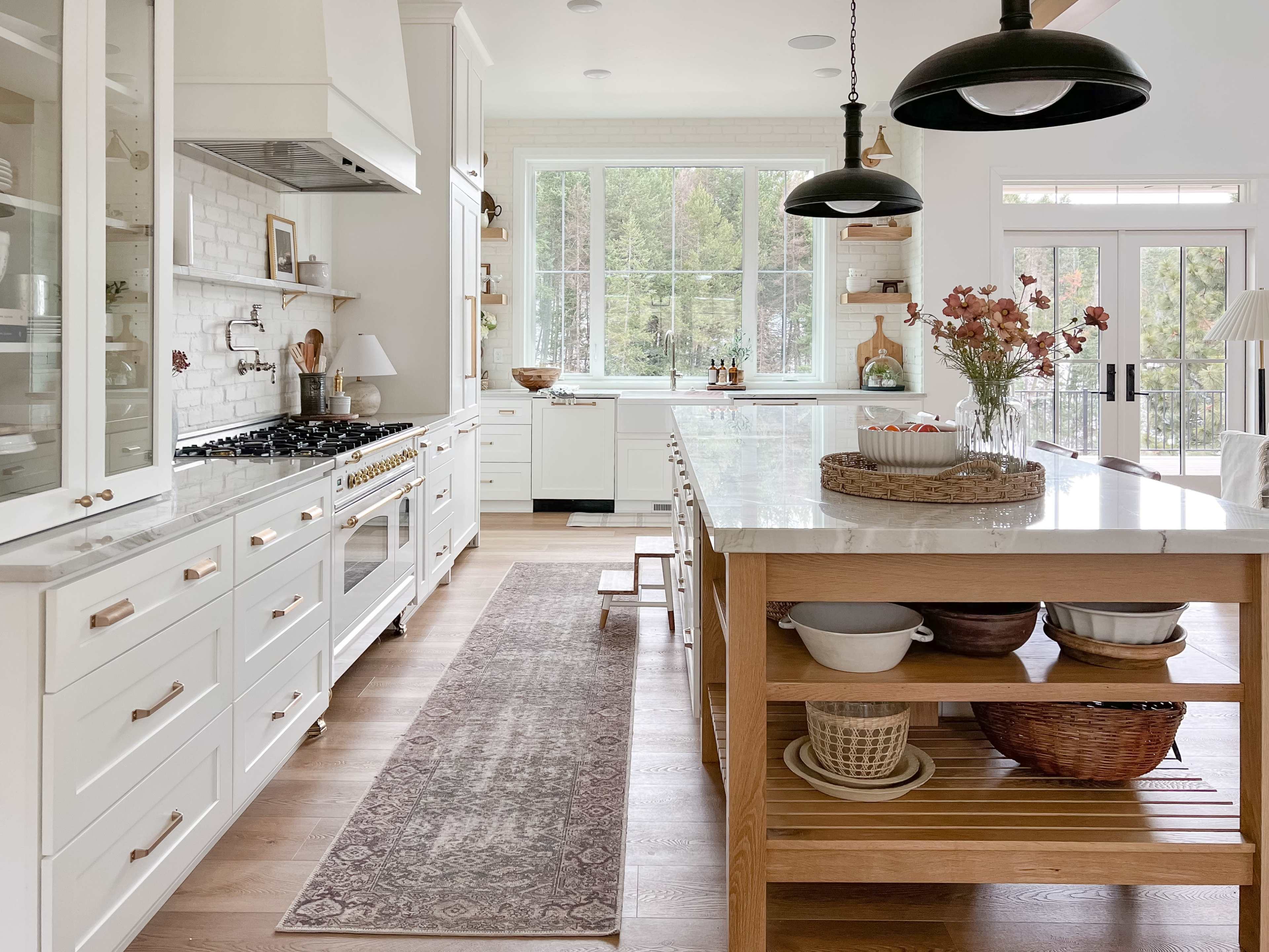 A modern kitchen features white cabinetry, a large island with wooden shelving, and a marble countertop, illuminated by hanging pendant lights.