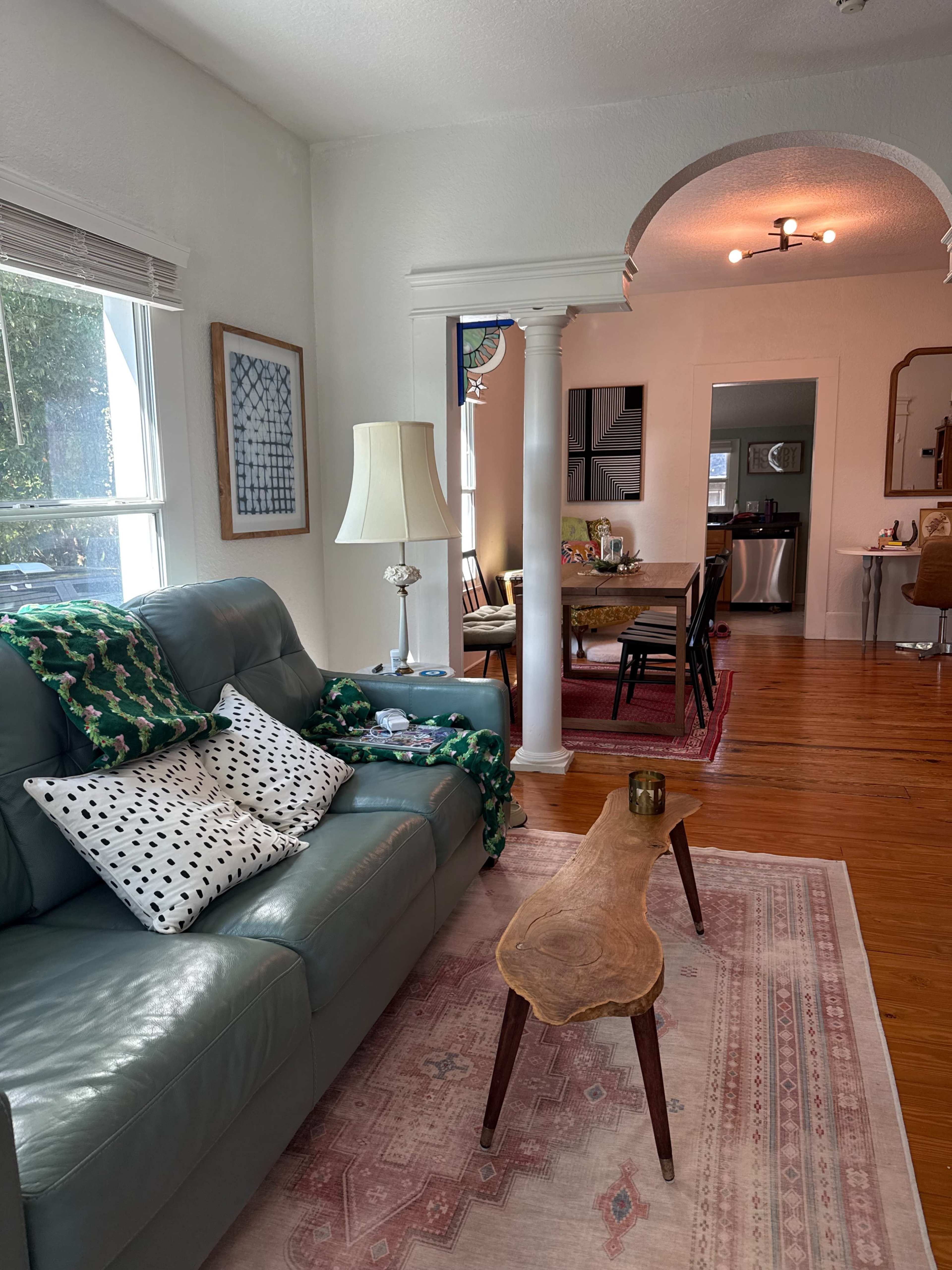 The image shows a cozy living room featuring a green couch with patterned pillows, a wooden coffee table, a rug, and an adjacent dining area visible through an archway.