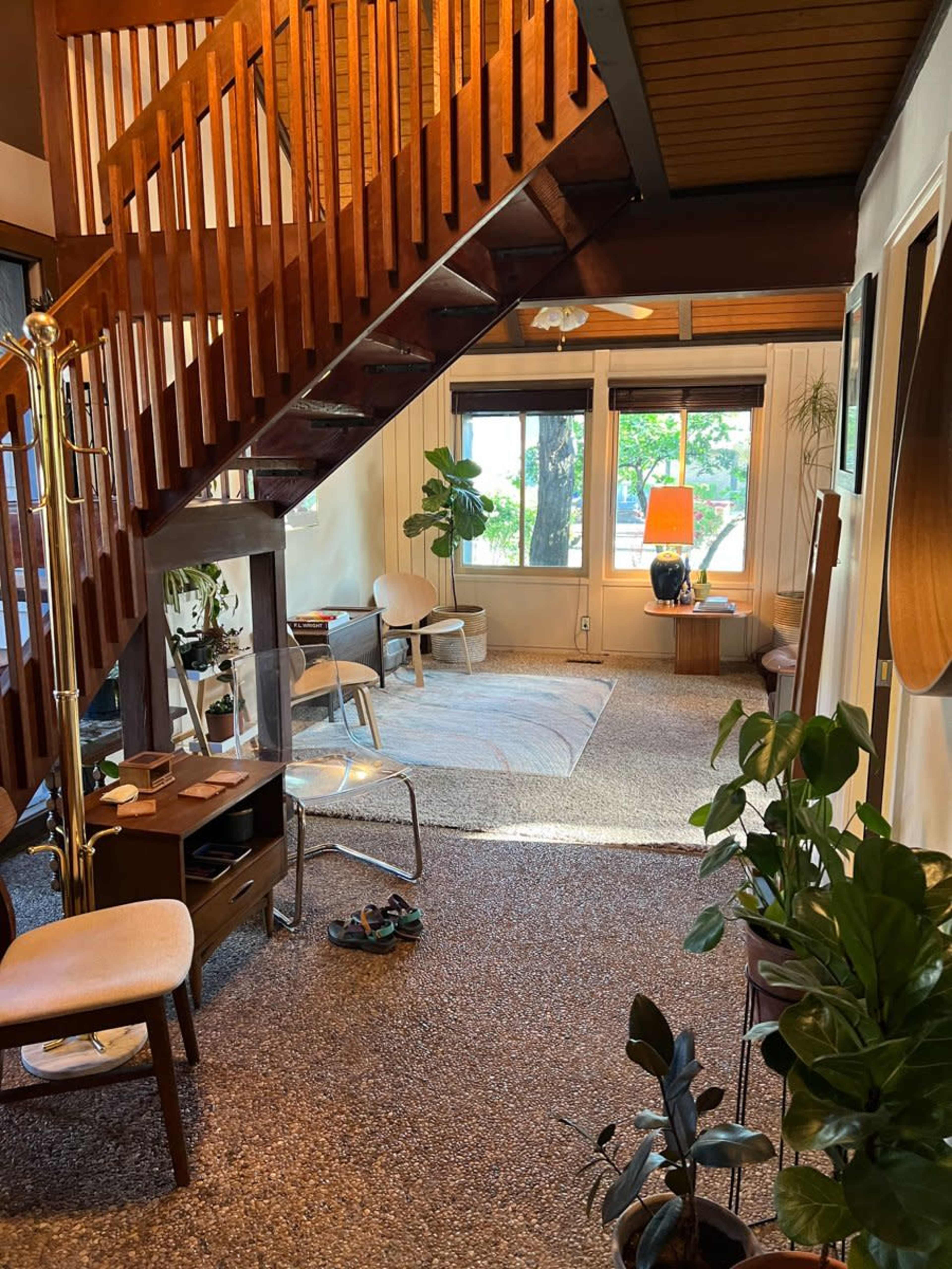 A cozy interior space featuring a staircase, seating area, plants, and a large window illuminated by natural light.