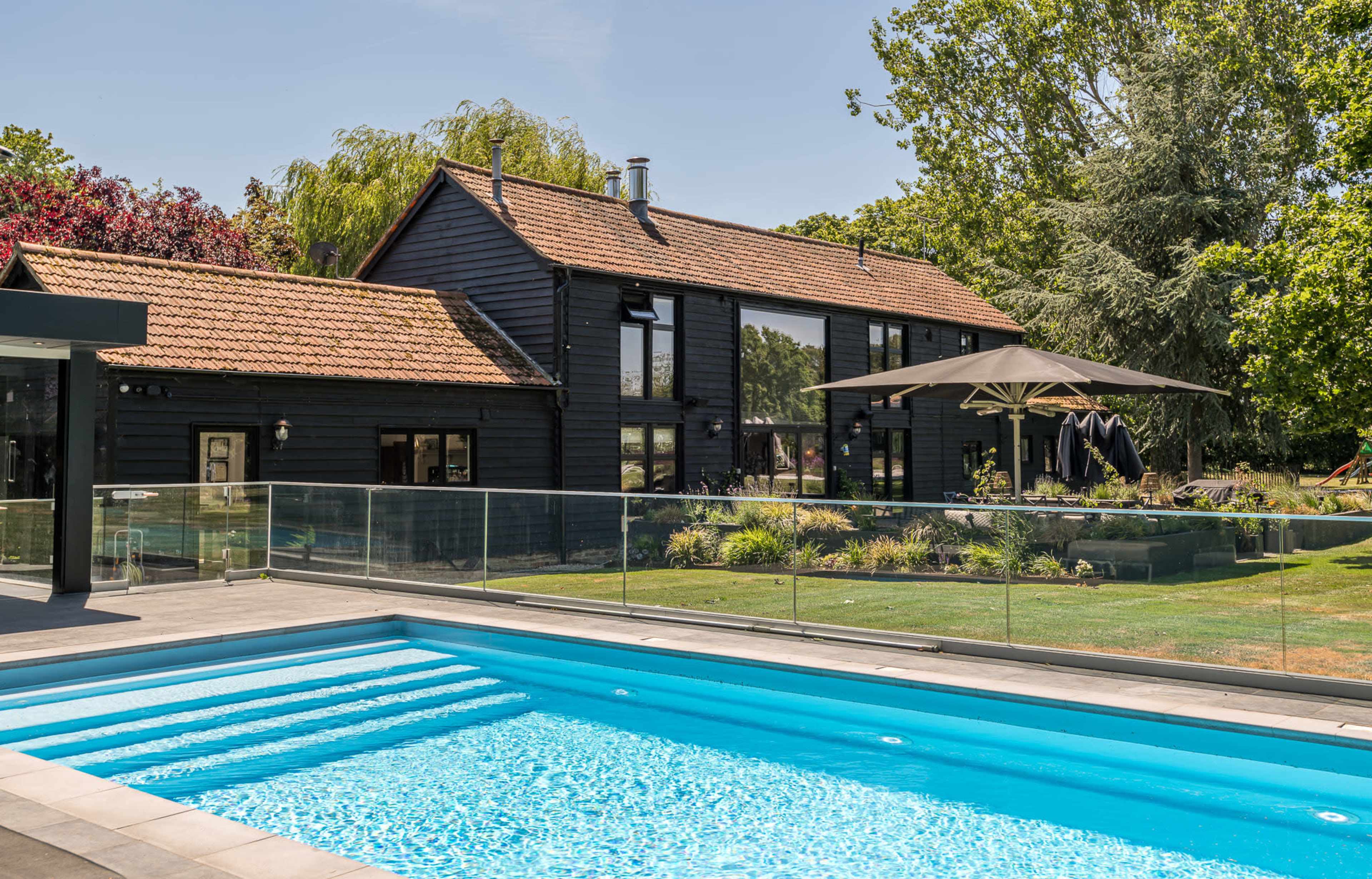 The image shows a modern black wooden house with large windows, surrounded by greenery and featuring a swimming pool in the foreground.