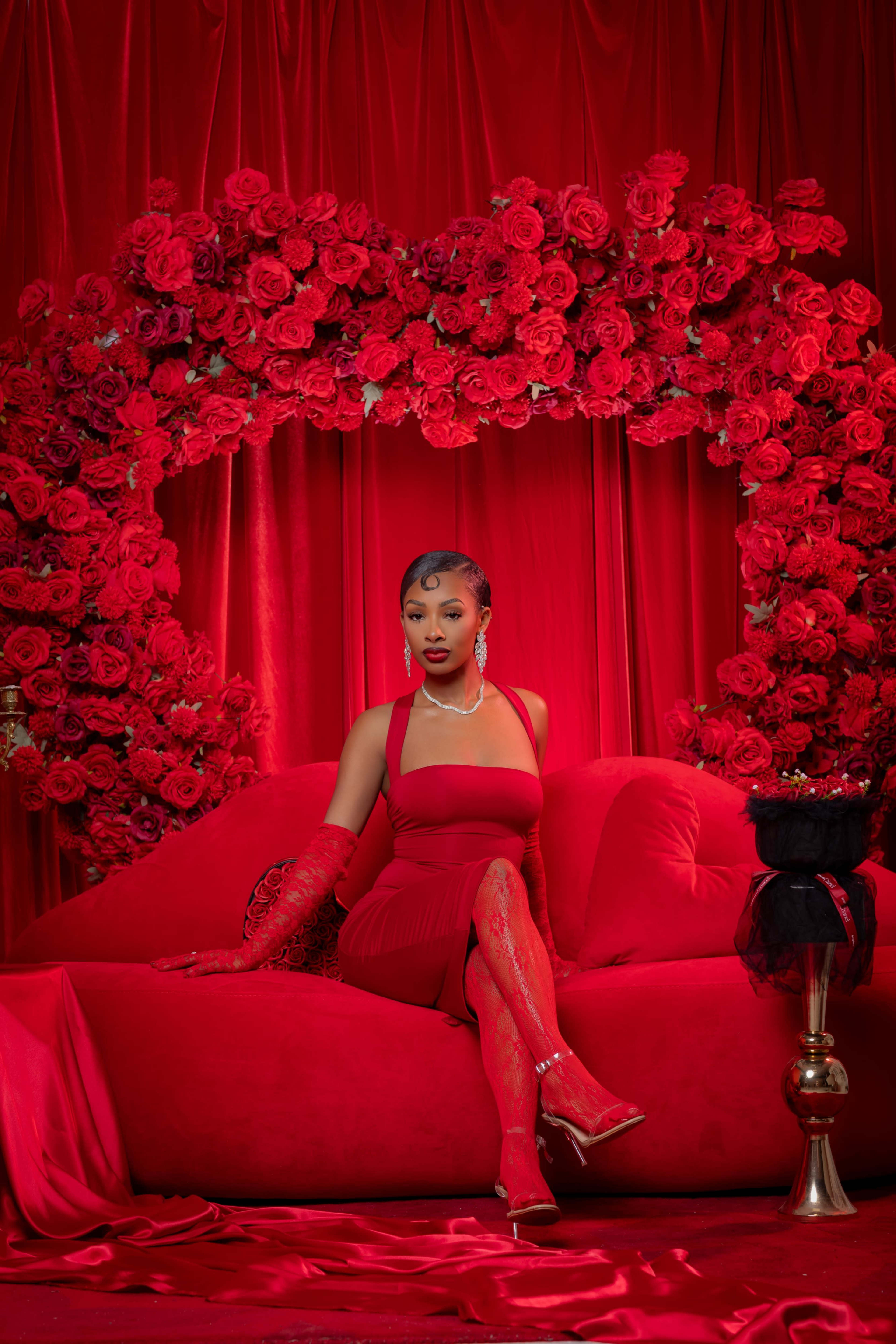 A woman sits elegantly on a red sofa surrounded by a large heart-shaped arrangement of red roses.