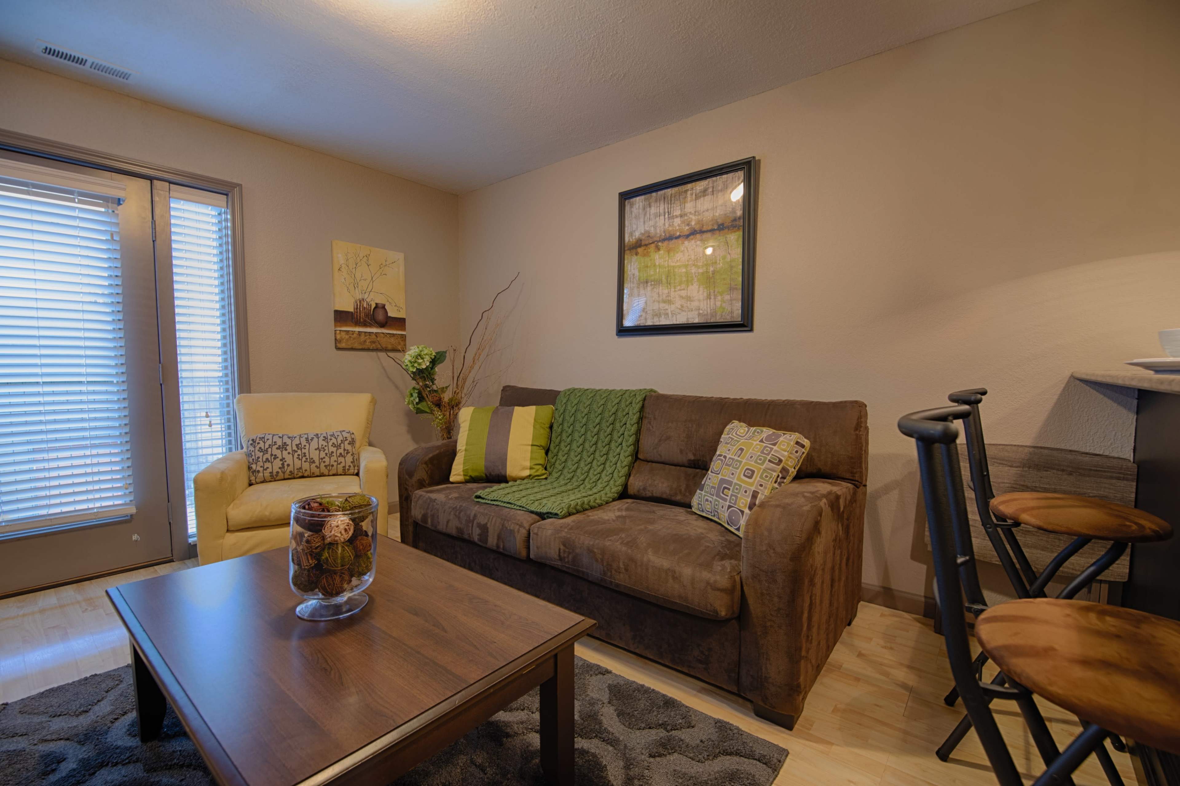 A living room with a brown couch adorned with green and patterned cushions, a wooden coffee table, and a small dining area featuring high stools.