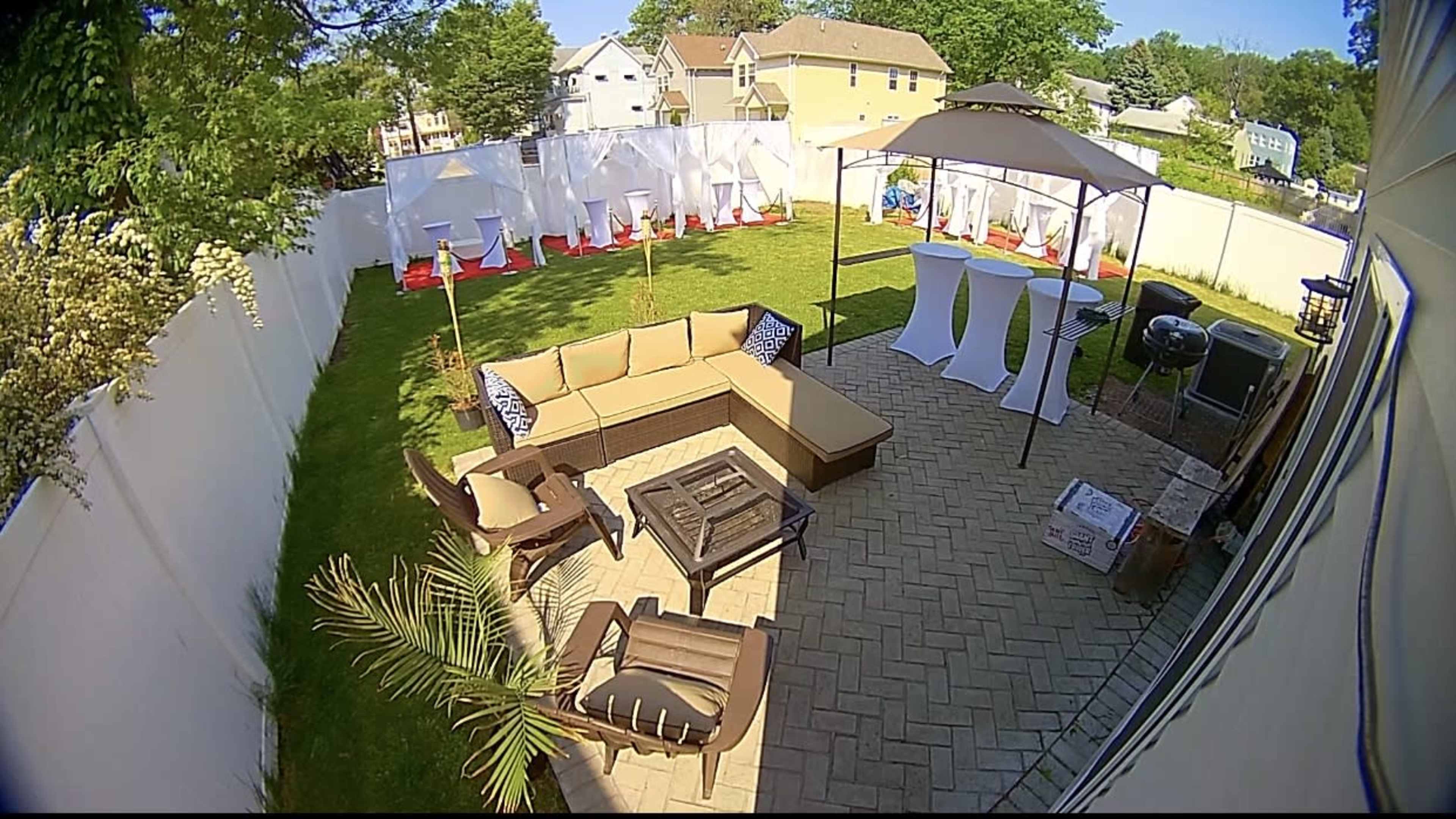 A backyard setup with a patio area featuring a sectional sofa, a fire pit, and several tables under a canopy, surrounded by a well-maintained lawn and white fencing.