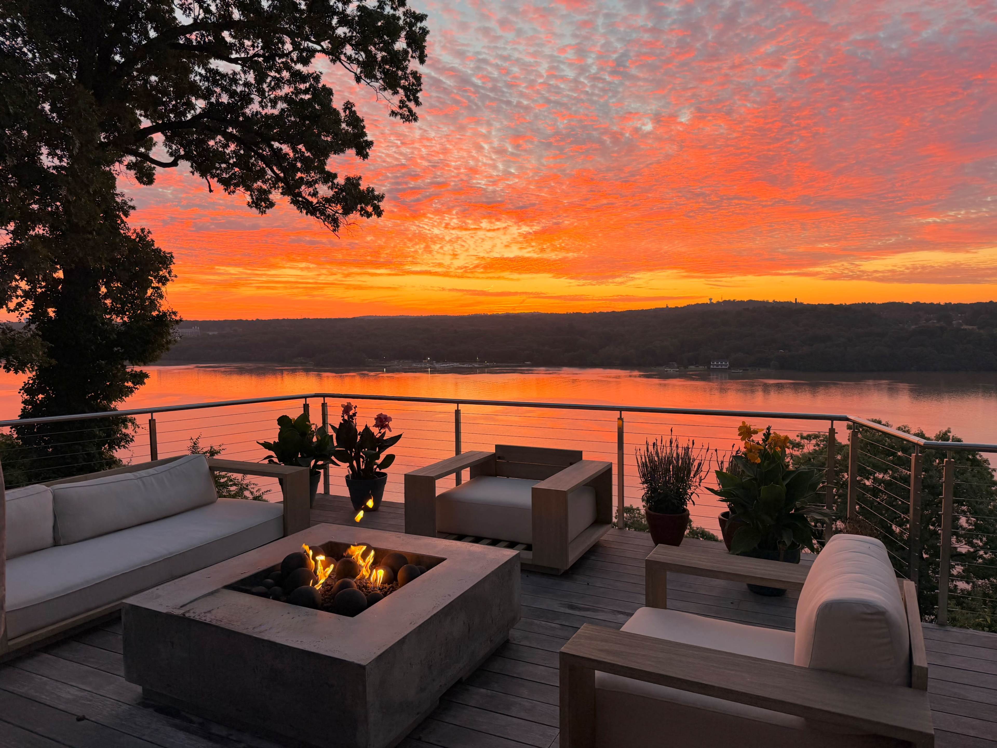 A wooden deck features modern outdoor furniture arranged around a fire pit, overlooking a river at sunset with vibrant orange and pink hues in the sky.