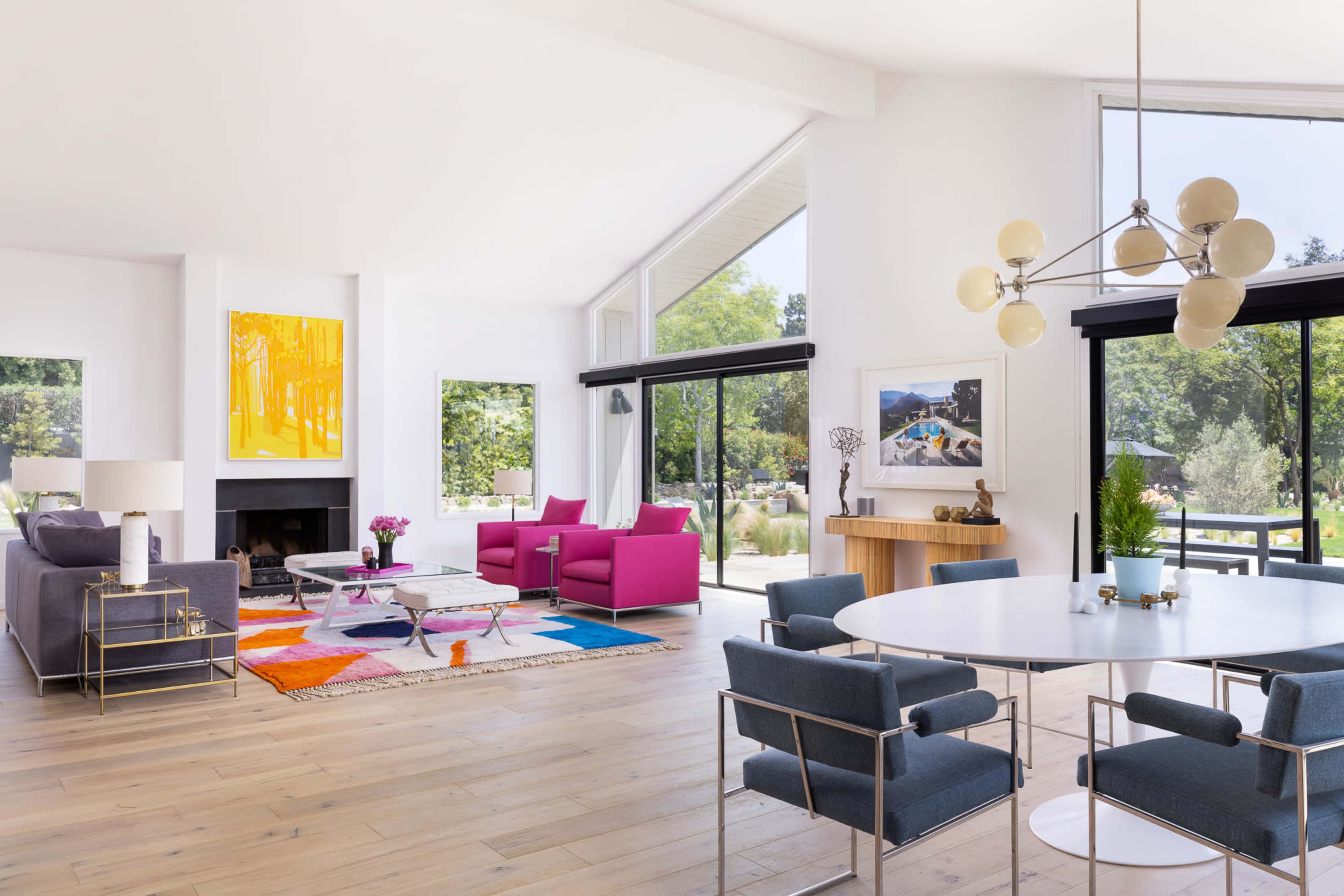 A bright, modern living room features colorful furniture, large windows overlooking greenery, and a mix of seating areas with a dining table.