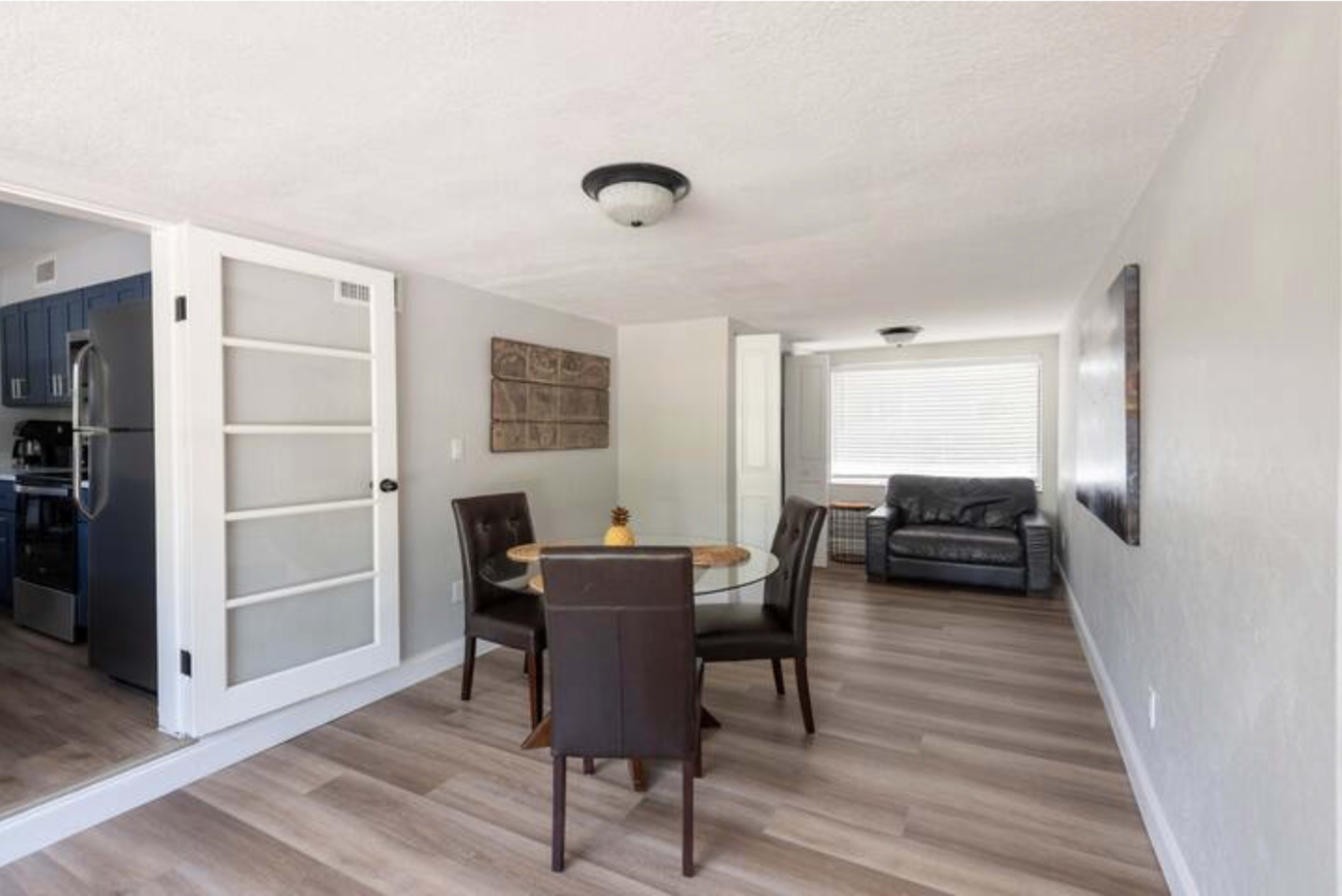 A small, modern dining area with a round glass table and four brown chairs, adjacent to a living room featuring a dark-colored couch.