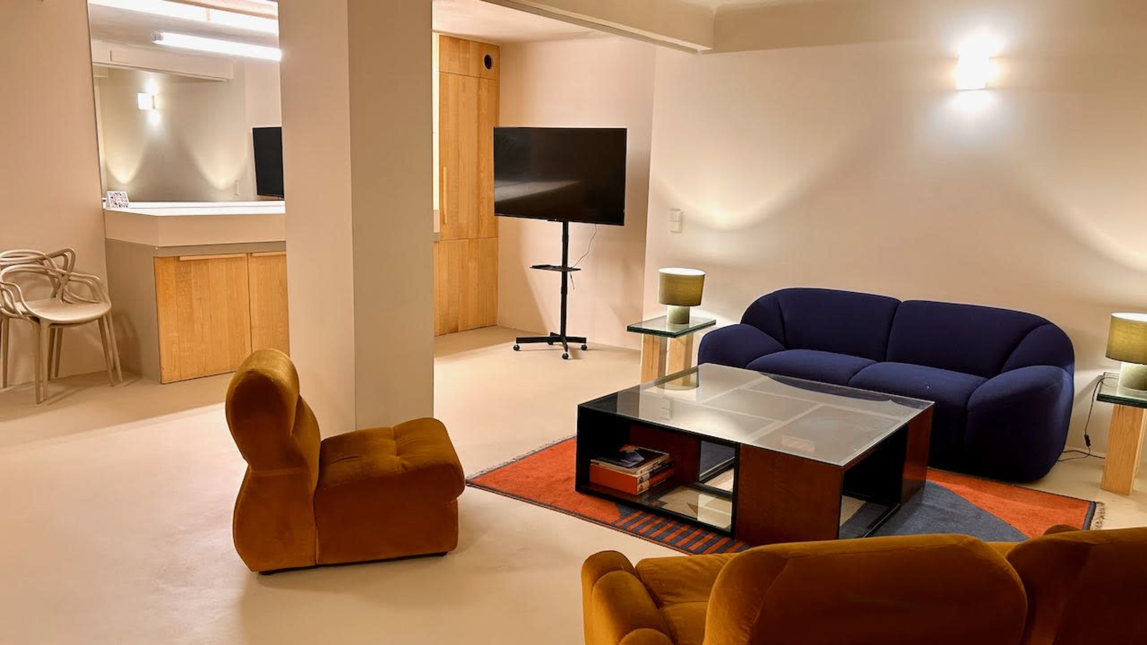 The image shows a modern living area featuring a blue sofa, two orange accent chairs, a glass coffee table, and a large TV on a stand against a light-colored wall.