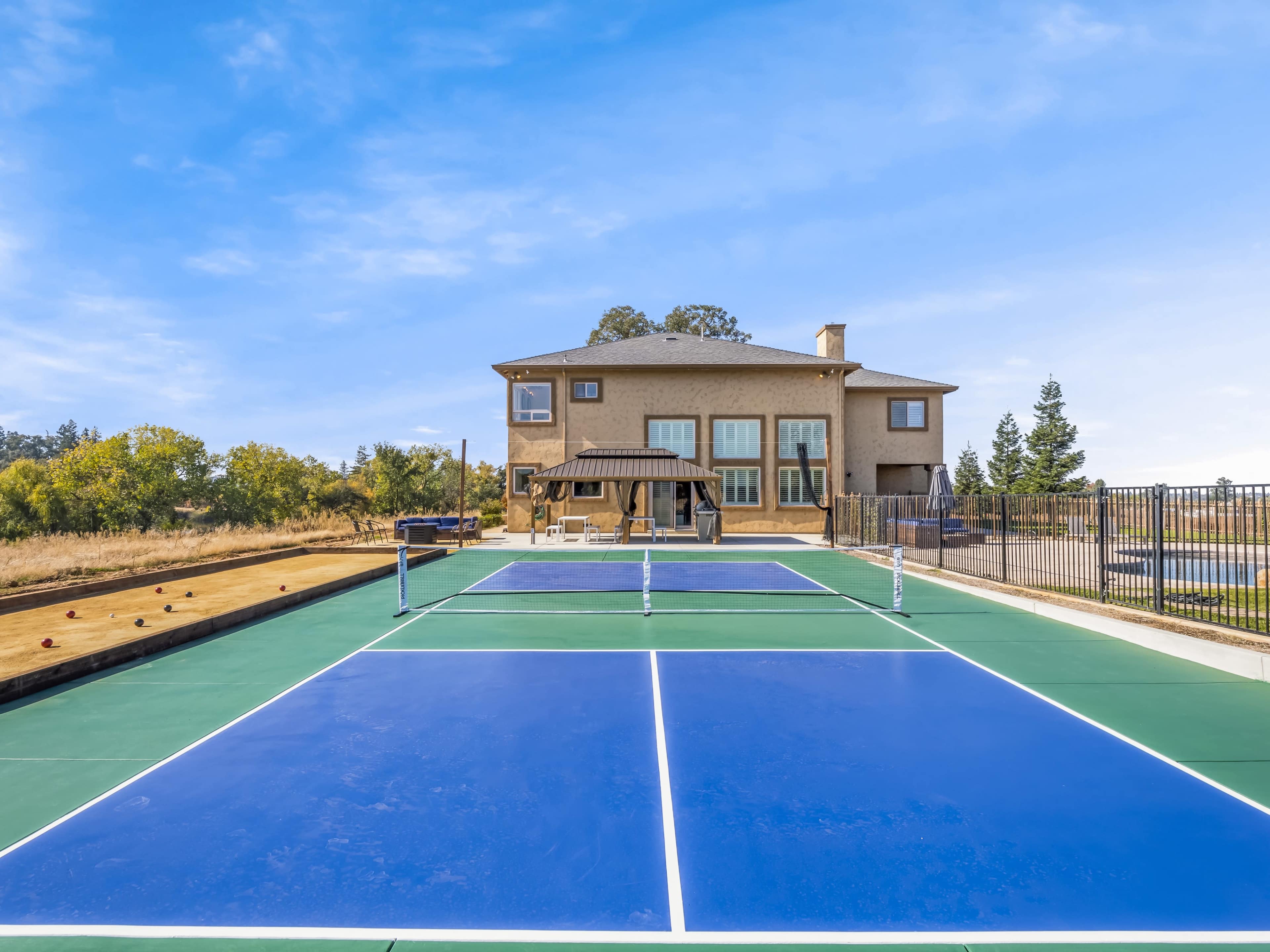 A tennis court with blue surfaces in the foreground, set against a large house and a clear blue sky in the background.