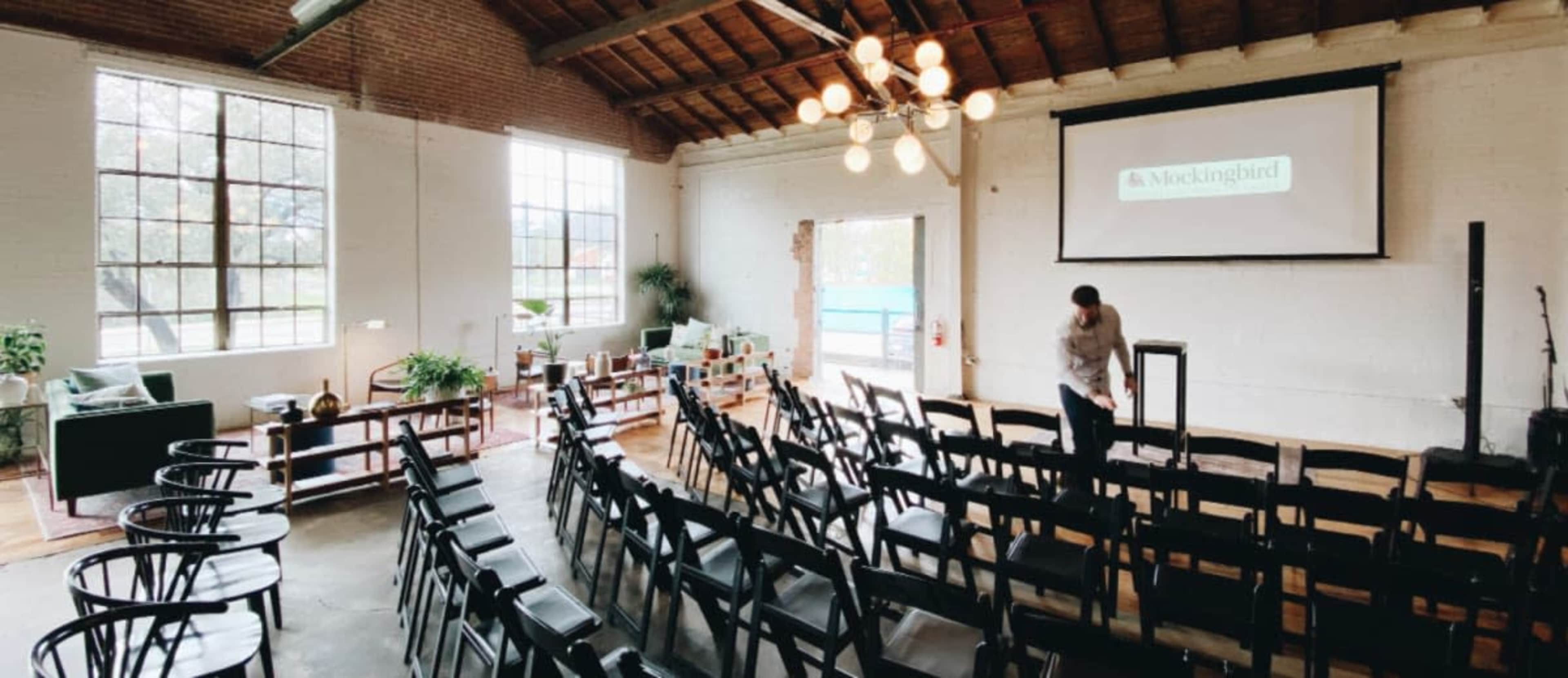 The image shows an industrial-style event space set up with rows of black chairs facing a screen, with large windows allowing natural light to fill the room.