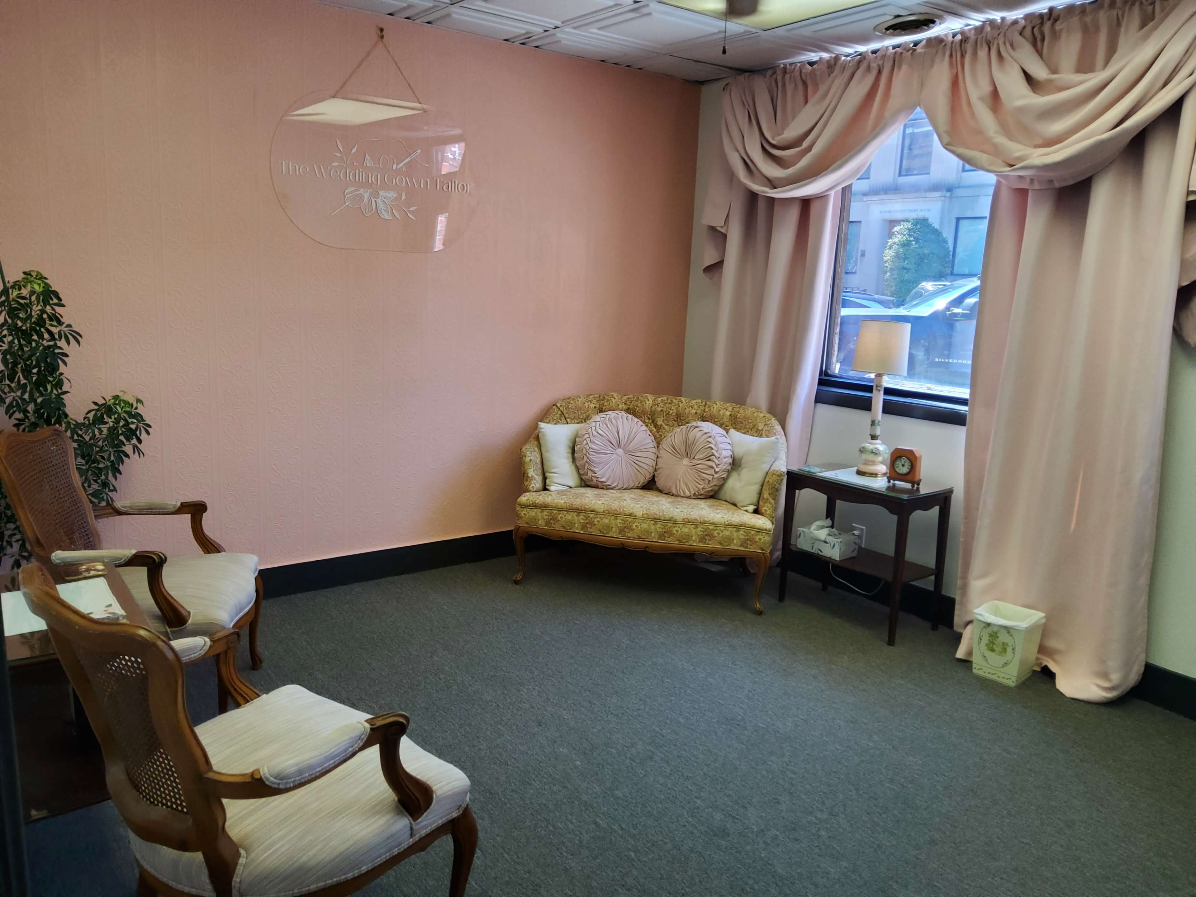 A waiting room with a pink wall, a patterned couch, two wooden chairs, a lamp, and a small table by a window.