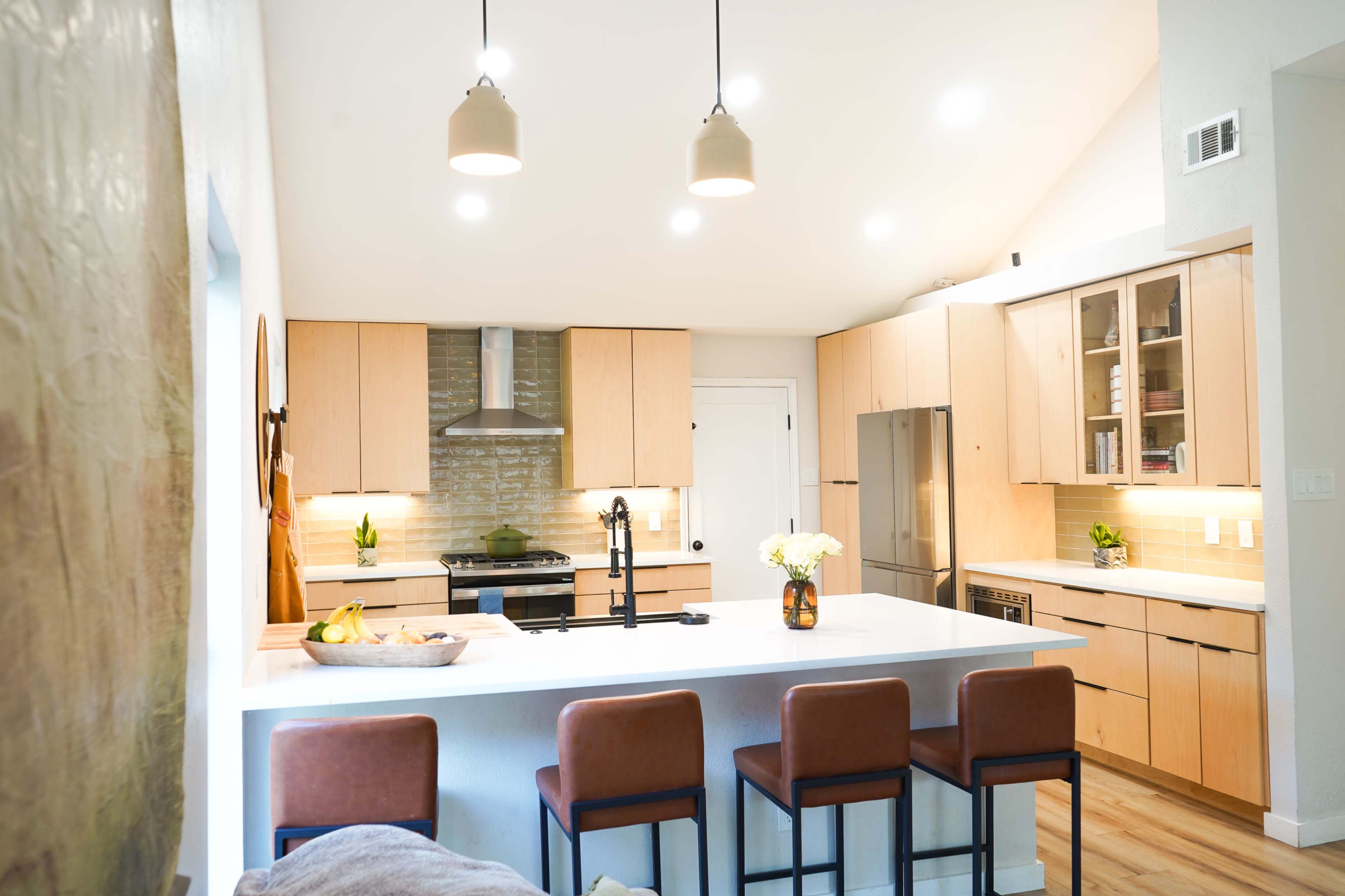 The image shows a modern kitchen with light wood cabinetry, a central island with bar stools, and stainless steel appliances.