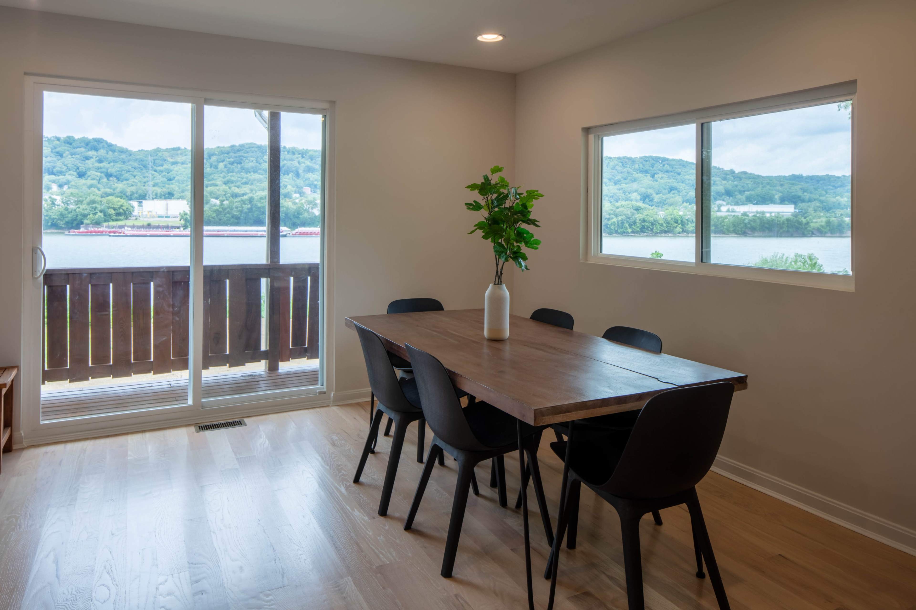A dining area with a wooden table and black chairs overlooks a scenic view of hills and a river through large windows.