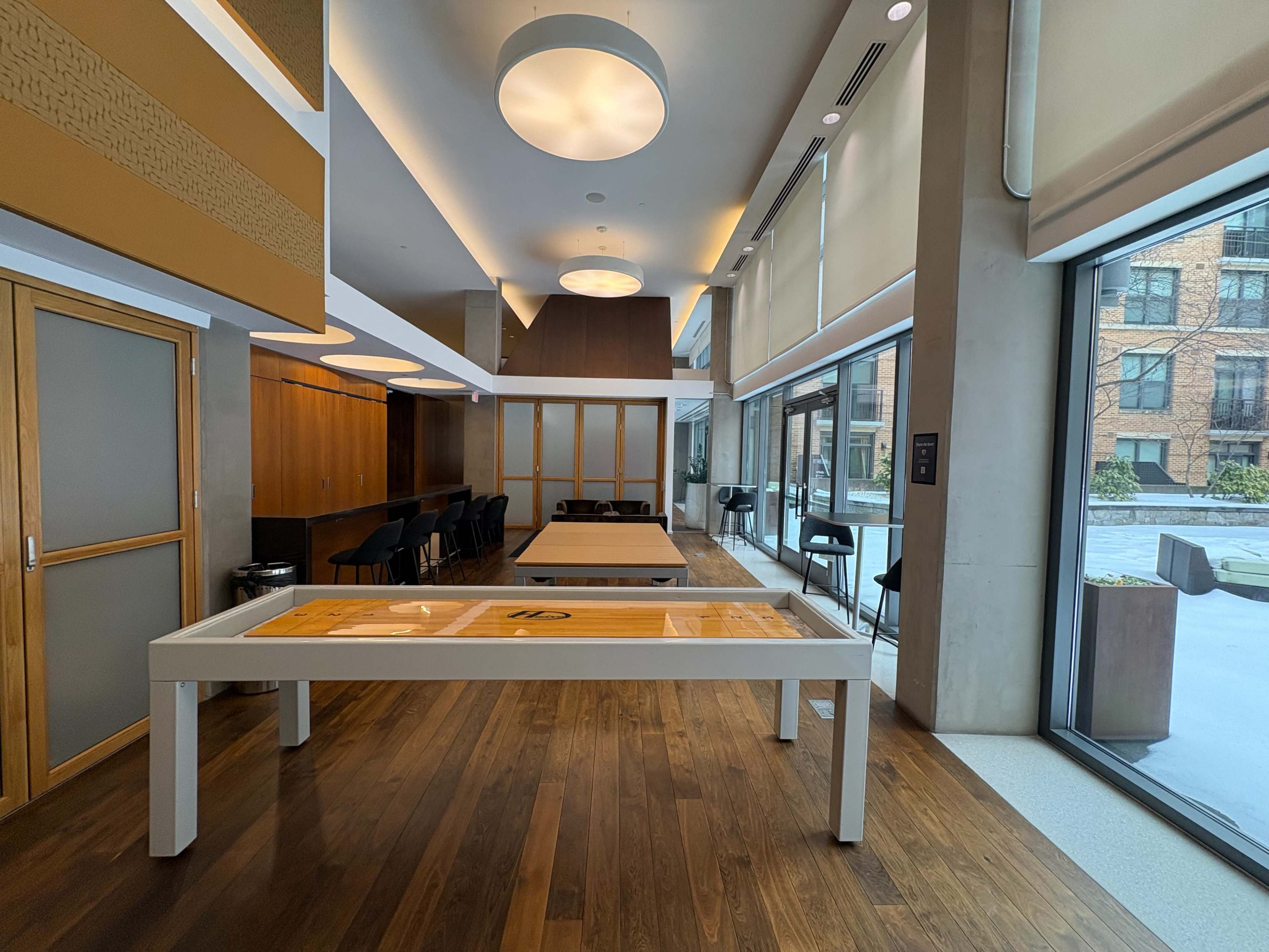 The image shows a modern, spacious interior featuring wooden flooring, contemporary furniture, and large windows overlooking an outdoor area.