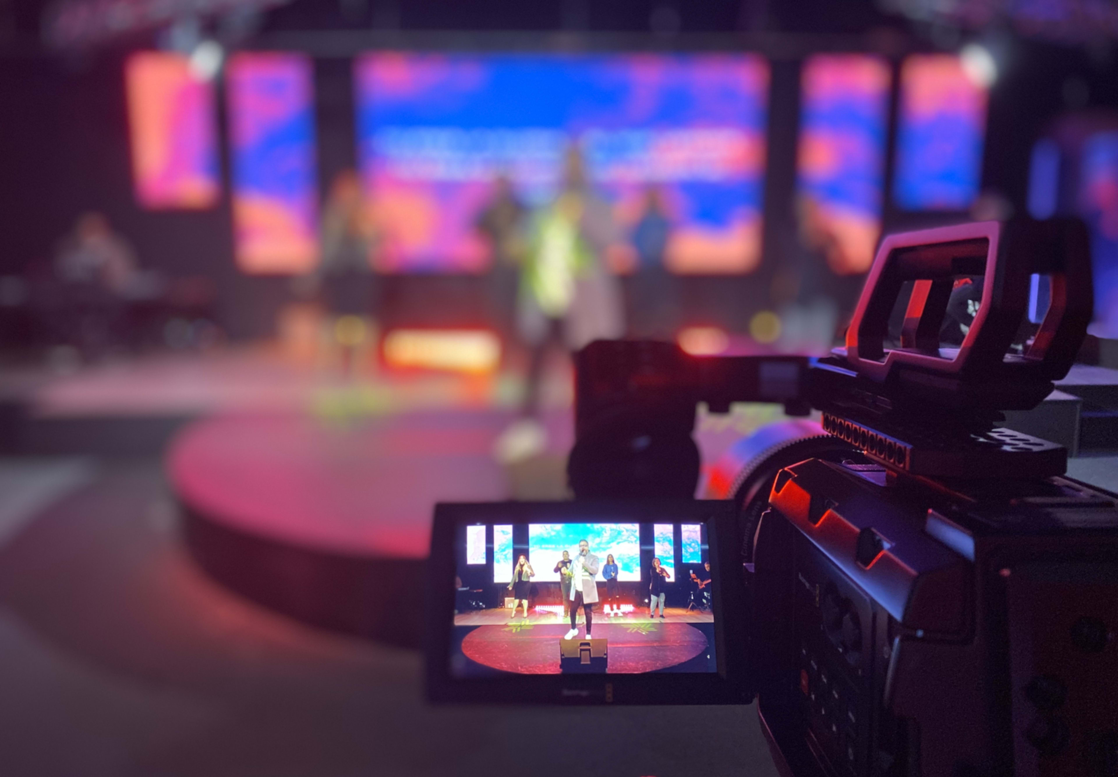 A video camera captures a performance on stage featuring multiple performers and a colorful backdrop.