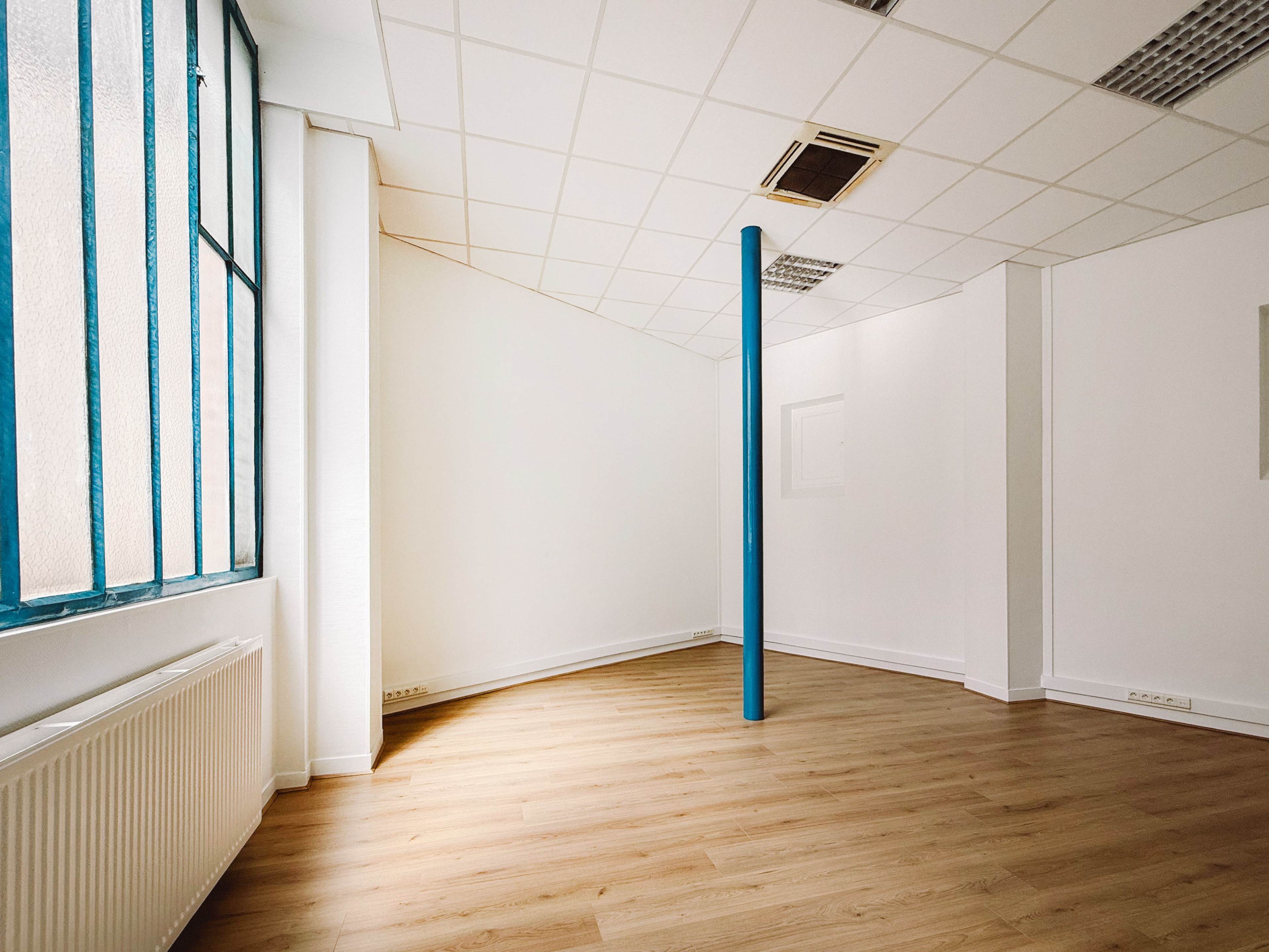 The image depicts a vacant room with a blue column in the corner, large windows allowing natural light, and light wooden flooring.