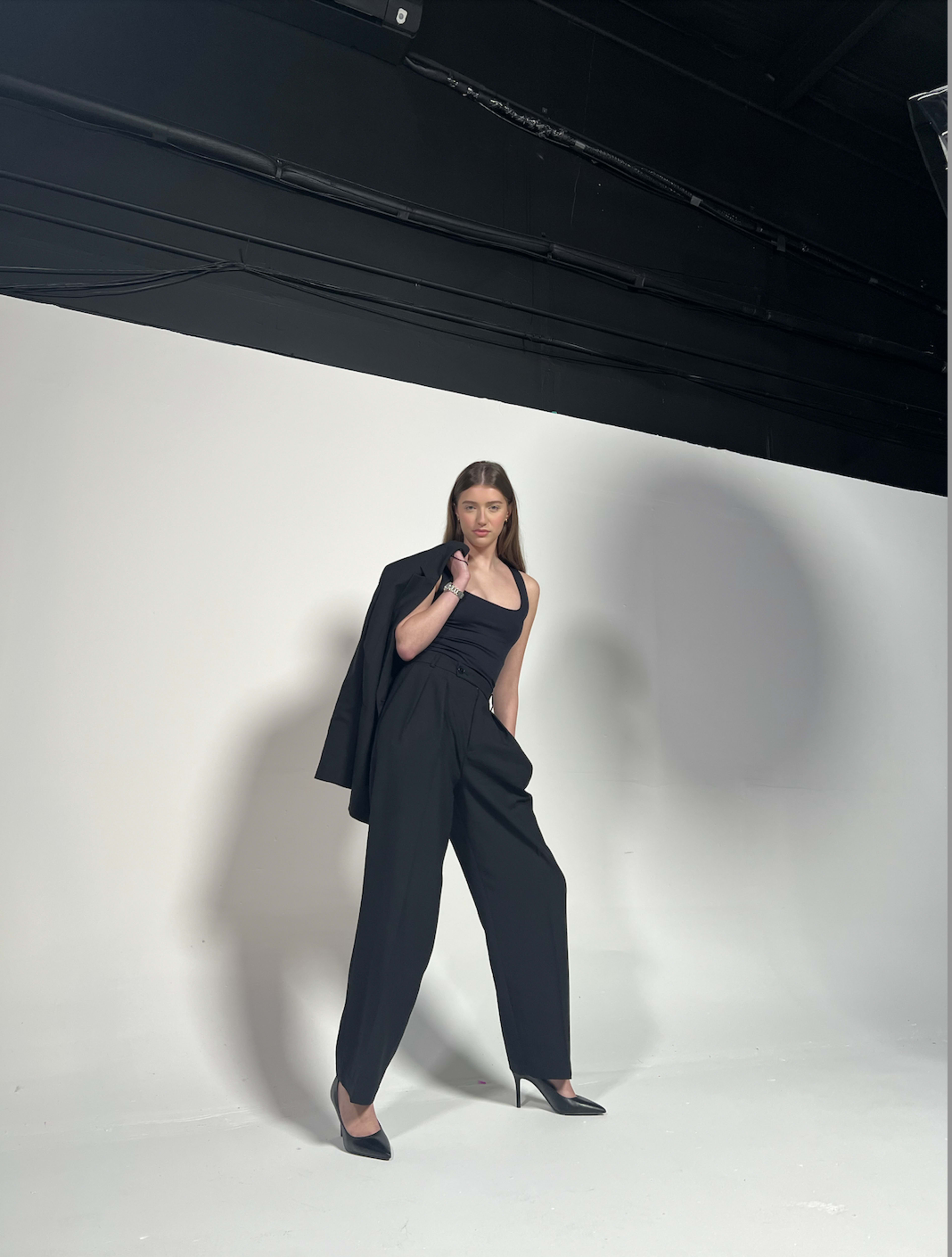 A model poses in a black outfit with a tailored blazer draped over one shoulder against a plain white background.